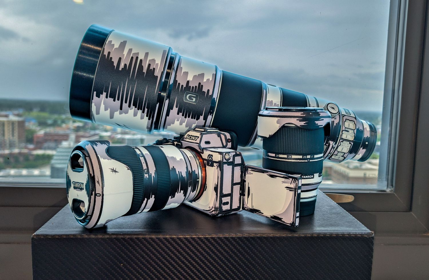 Two camera lenses with custom cityscape skin wraps displayed on a dark surface near a window with city views.