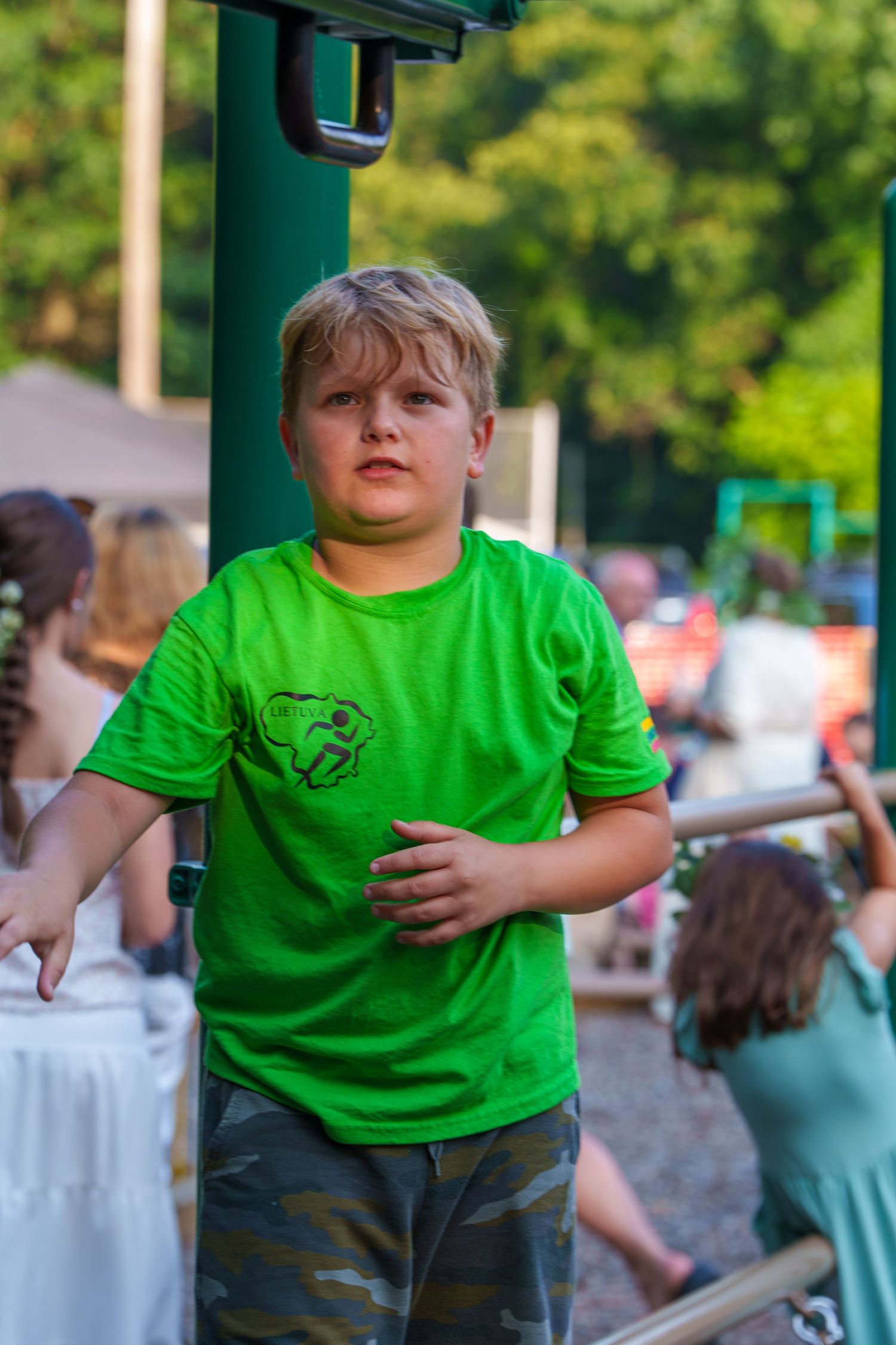 Young boy in bright green t-shirt and camo shorts playing on outdoor playground equipment.