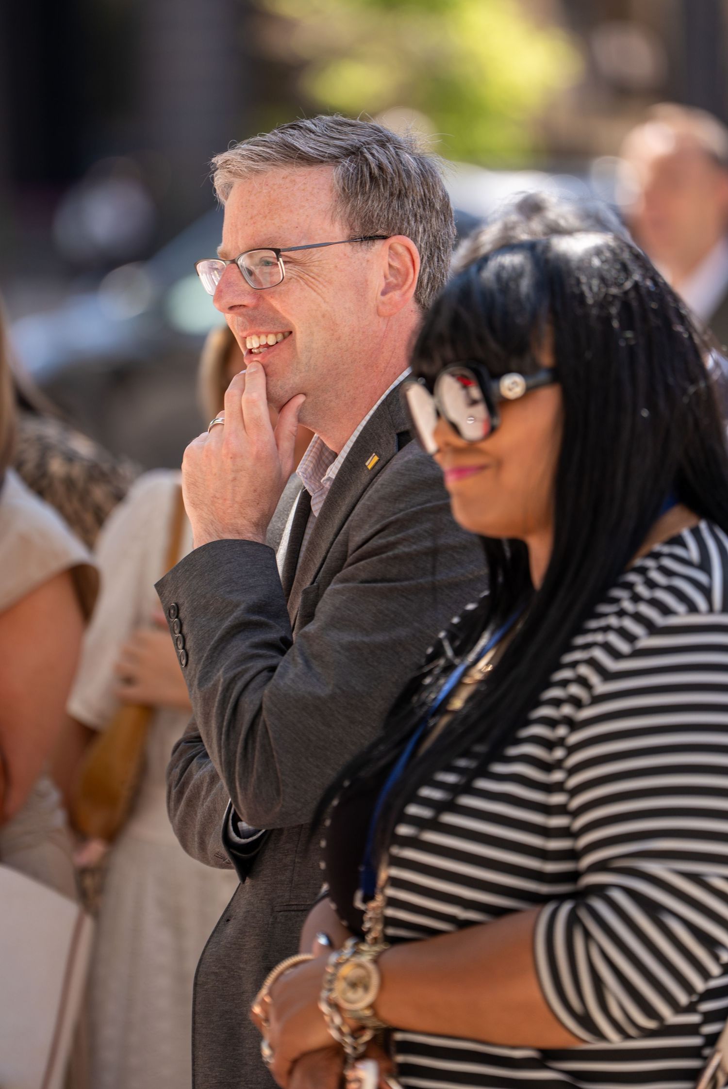 Man in gray suit smiling with hand on chin, woman in striped shirt wearing sunglasses at outdoor event.