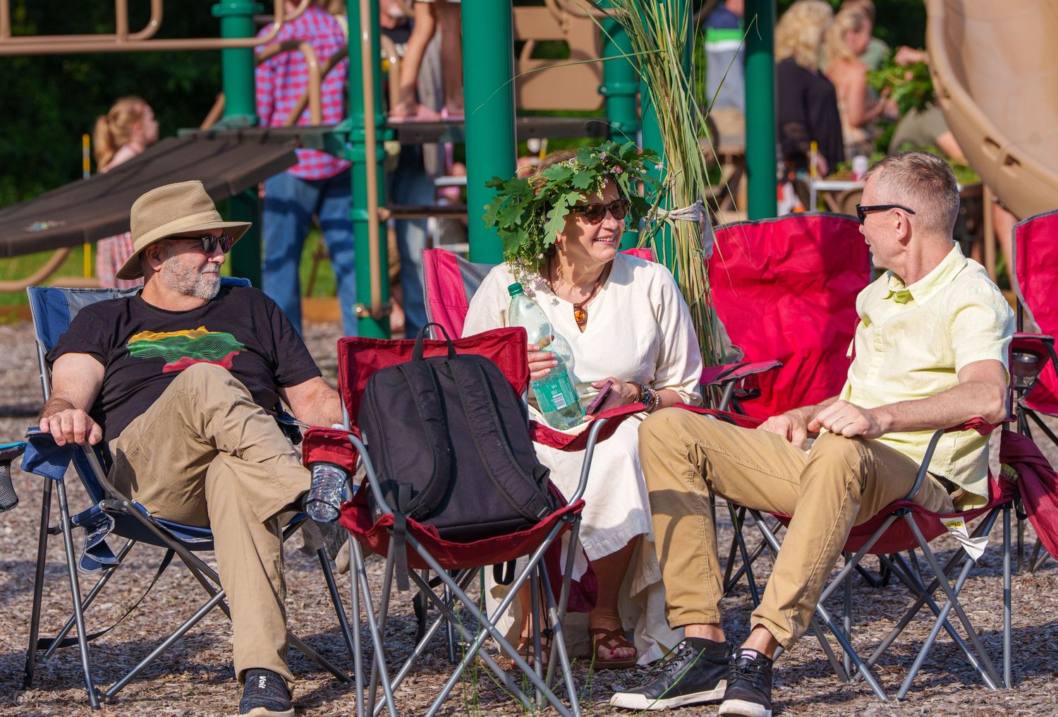 Three adults relaxing in folding chairs outdoors, one wearing a floral wreath at a summer festival.