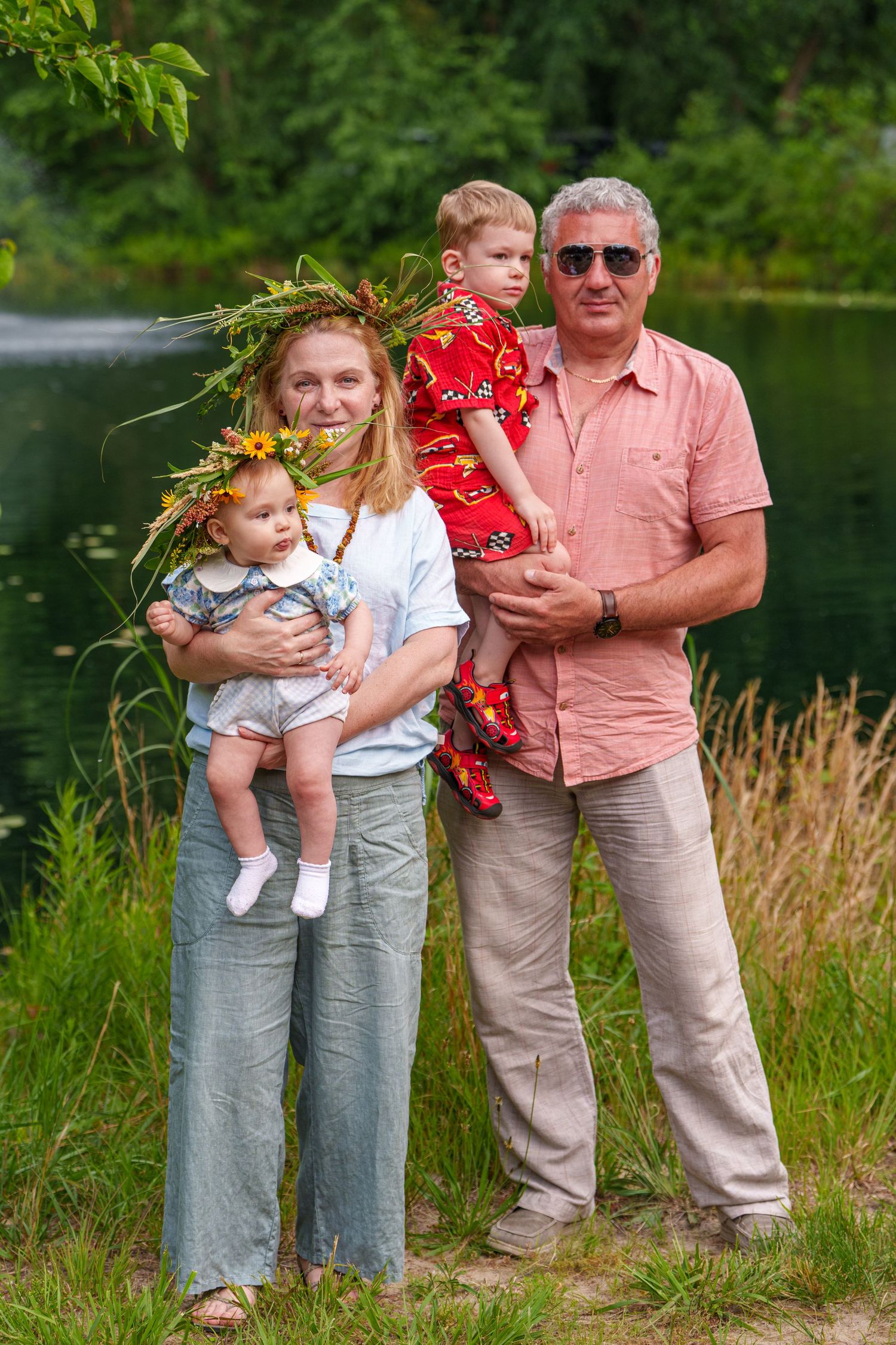 Family portrait outdoors by a pond, woman with floral crown holding baby, older man holding toddler in red outfit.