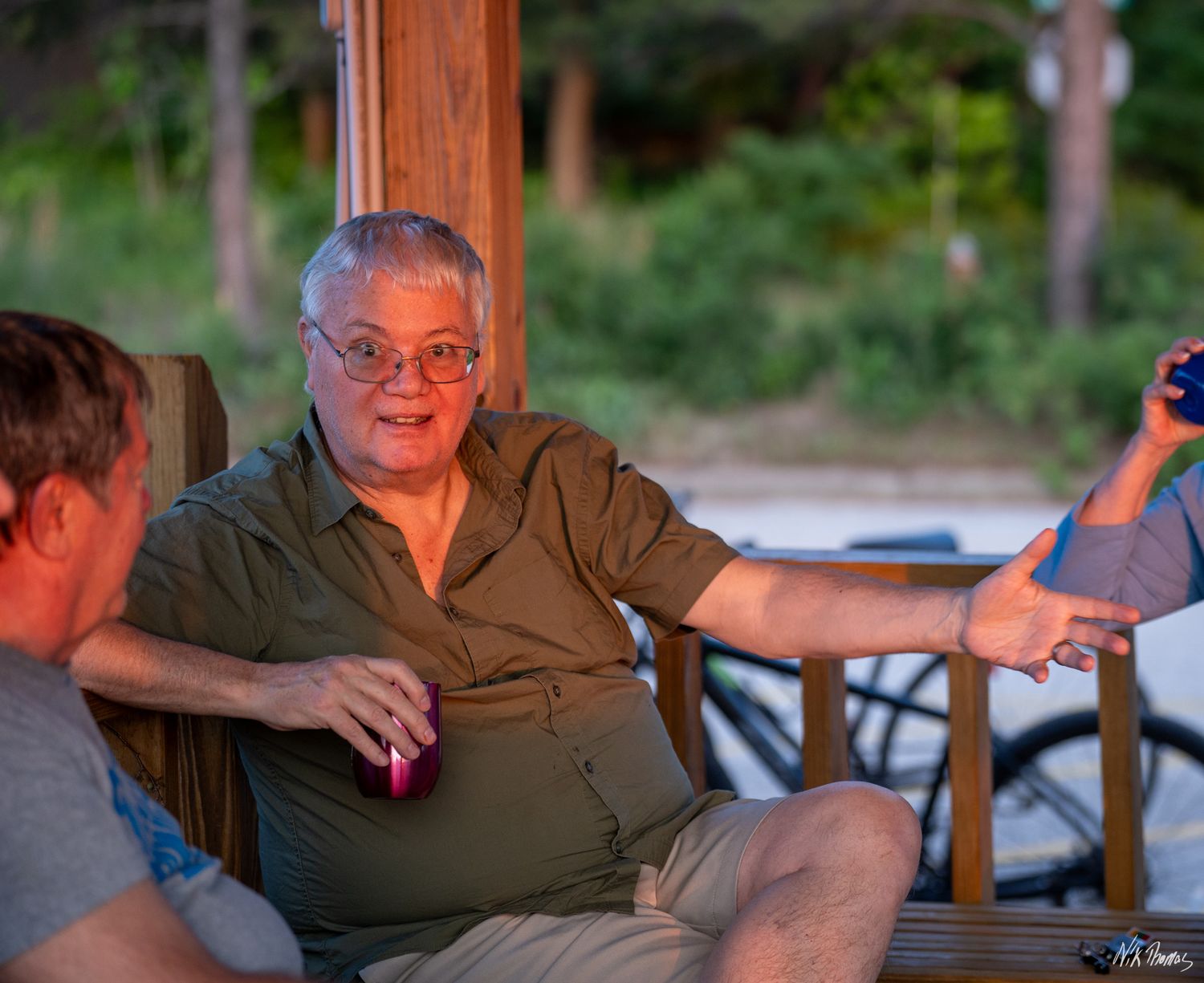 An older man wearing glasses and a green shirt socializes outdoors on a porch at dusk.