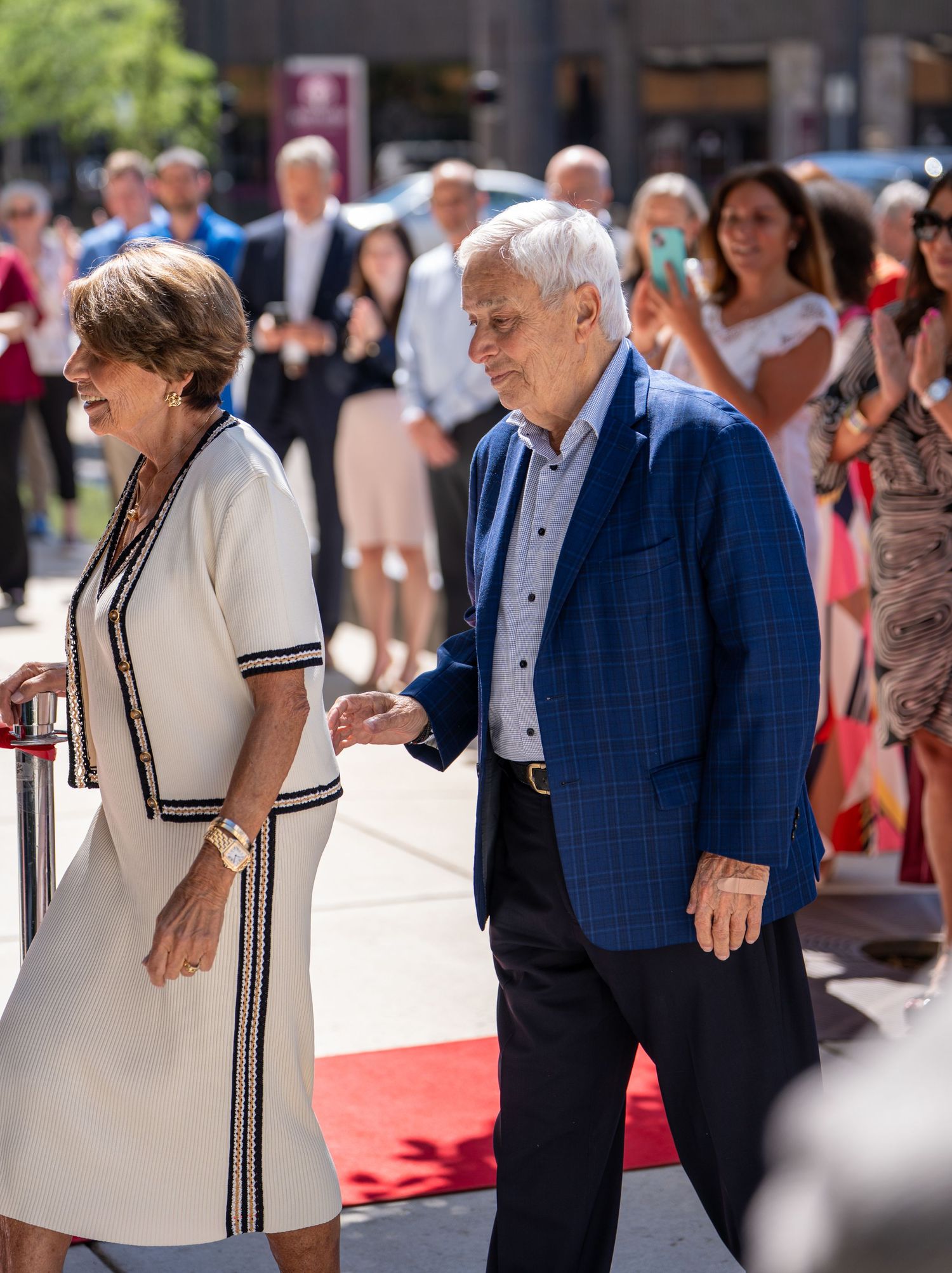 An elegant older couple walks a red carpet at an outdoor event, woman in white dress, man in blue blazer.