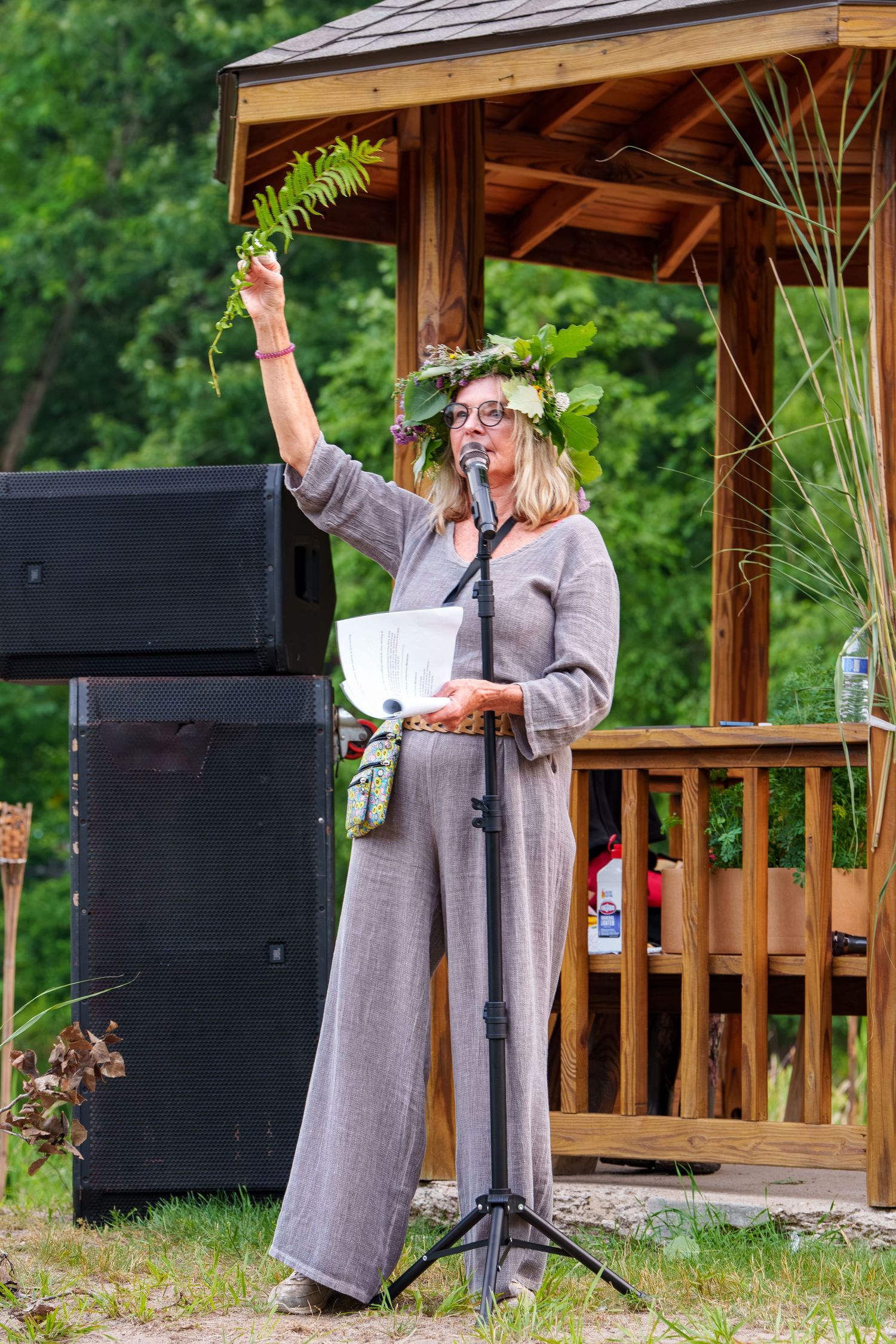 Woman in floral crown speaking at microphone podium at an outdoor cultural ceremony.