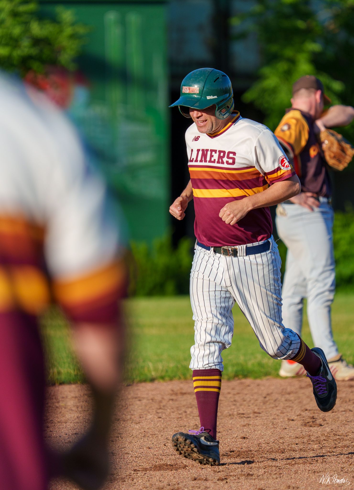Baseball player in Liners uniform running bases wearing green helmet during a sunny outdoor game.