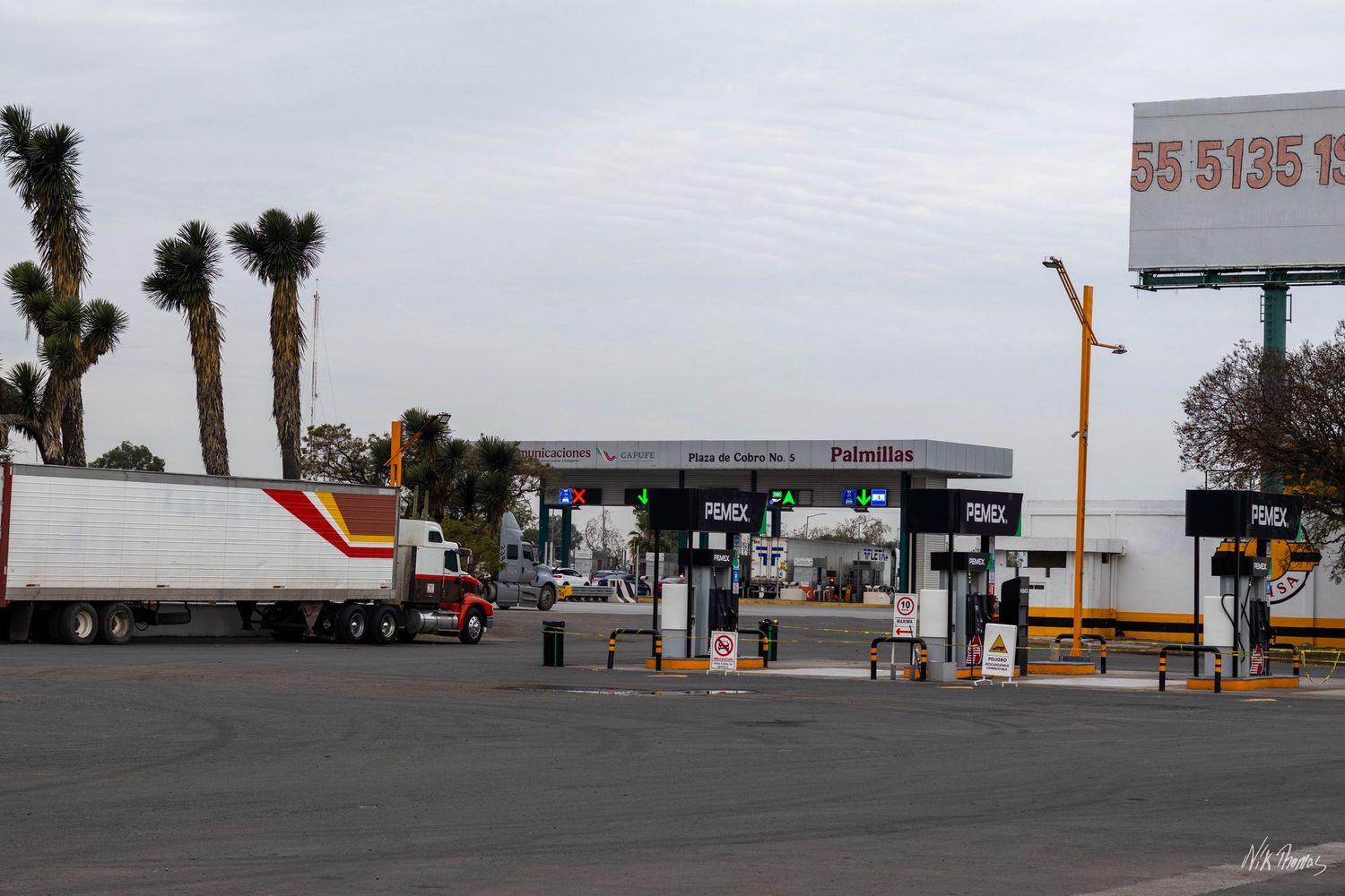 Semi-truck at a Mexican toll plaza with palm trees and a billboard displaying a phone number.