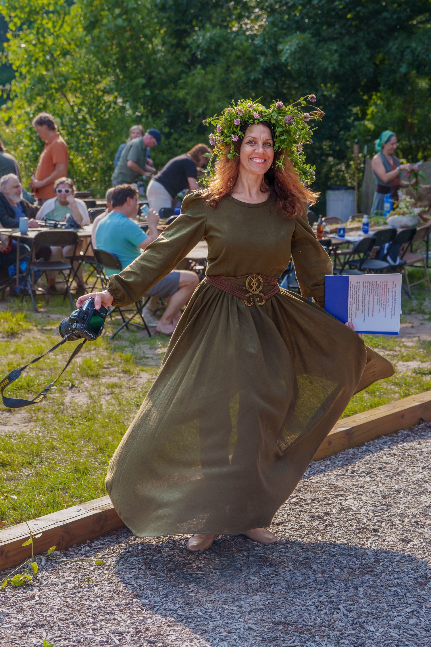 Smiling woman in olive green medieval dress with leaf crown twirls at outdoor festival.