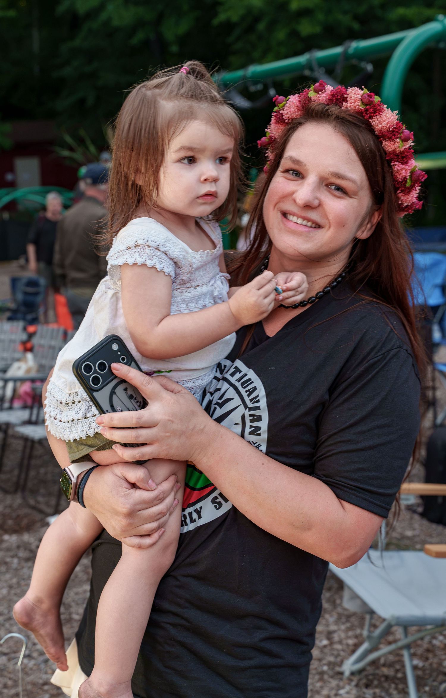 Smiling woman wearing a pink flower crown holds a young toddler girl at an outdoor event.