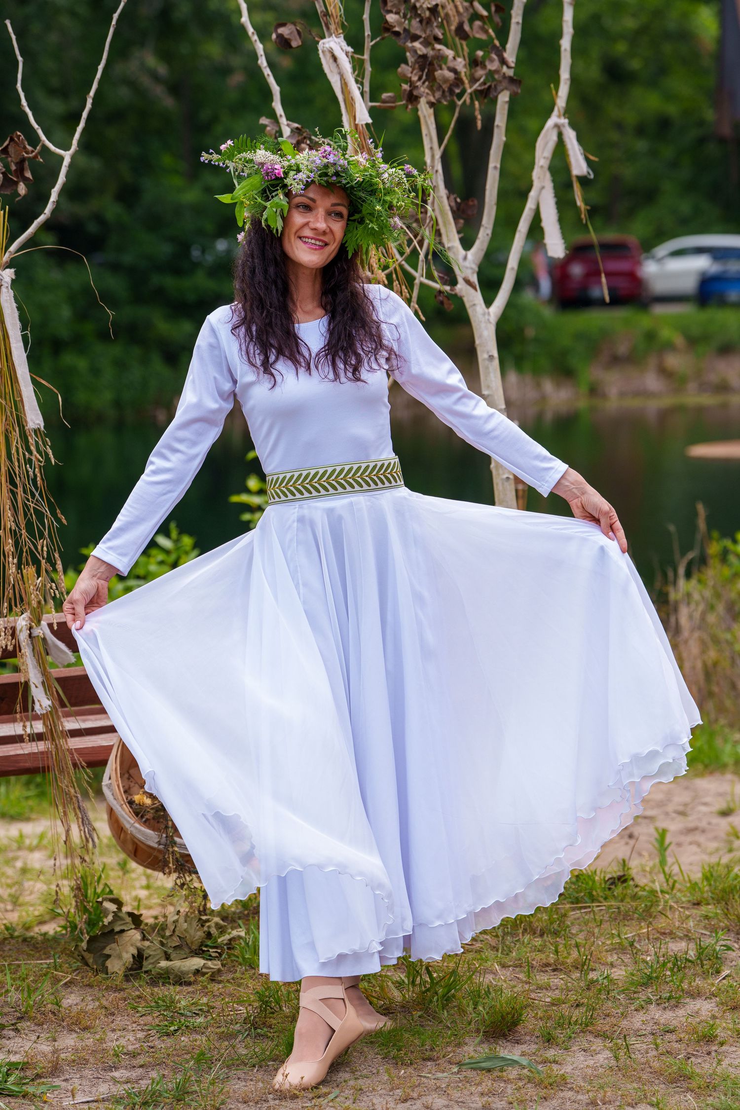Woman in white dress and floral crown twirls gracefully near a serene woodland lake.