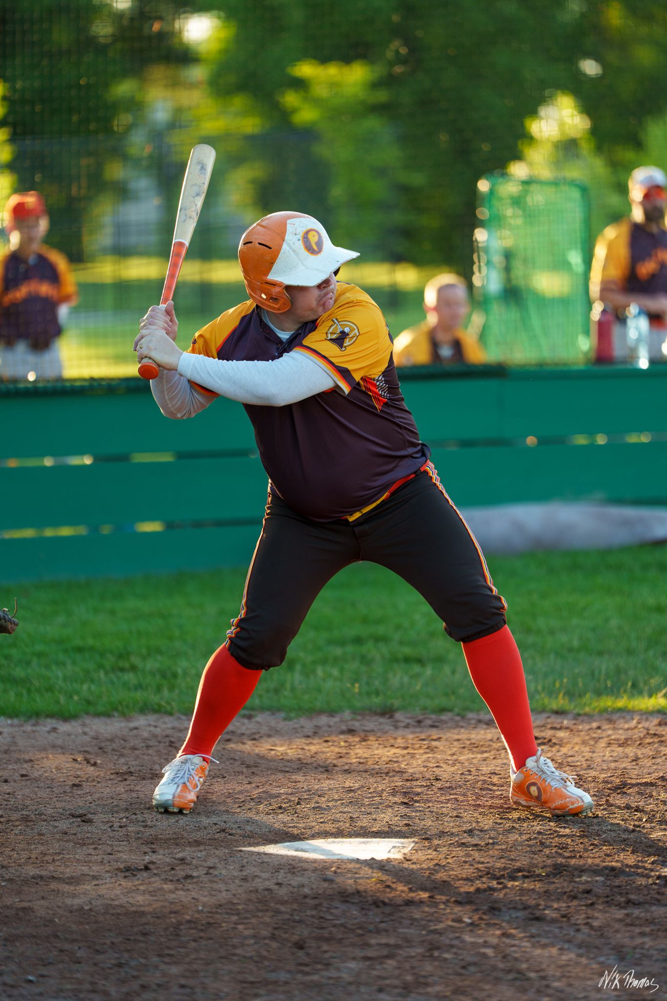 Softball batter in orange and brown uniform ready to swing at home plate during an outdoor game.