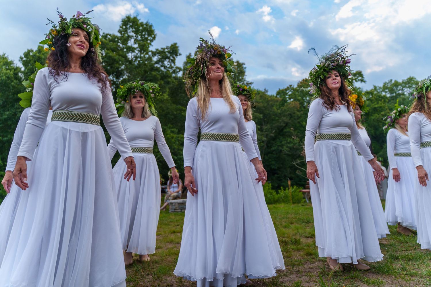 Women in white dresses and floral wreaths performing a traditional Lithuanian folk dance outdoors.