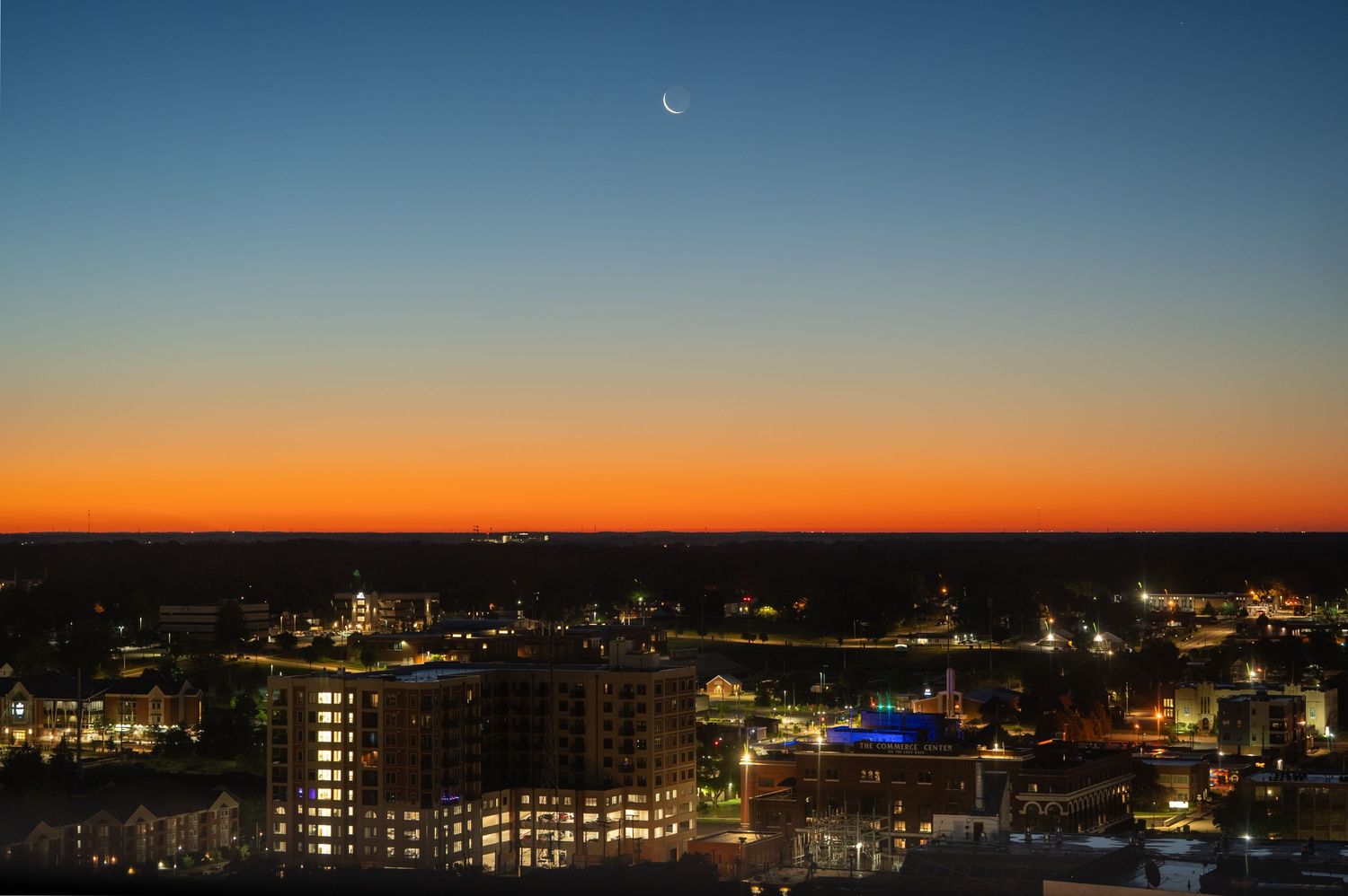 Aerial cityscape at twilight with crescent moon above glowing orange sunset horizon over illuminated urban skyline.