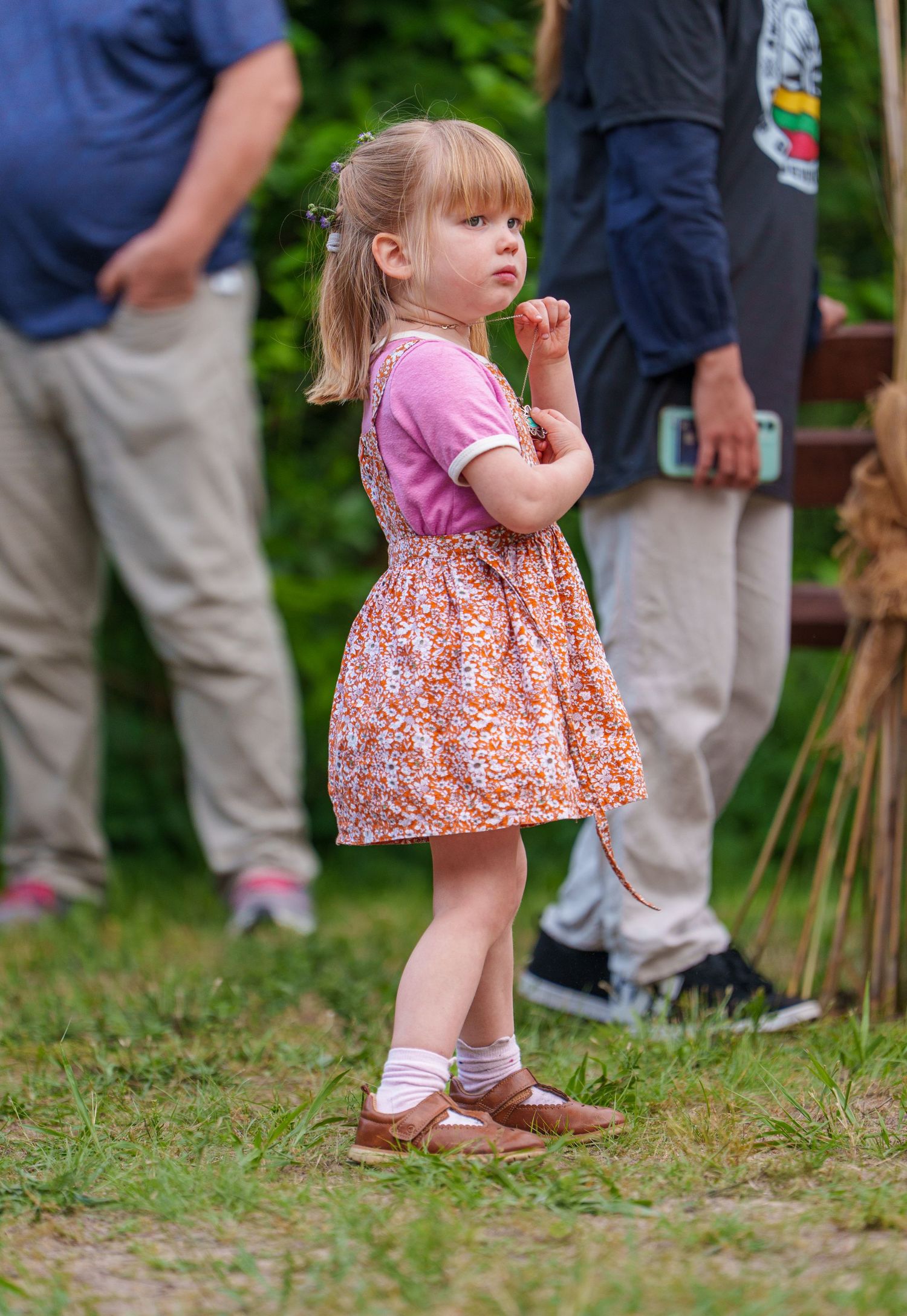 Adorable toddler girl in floral dress and pink shirt standing on grass at outdoor event.