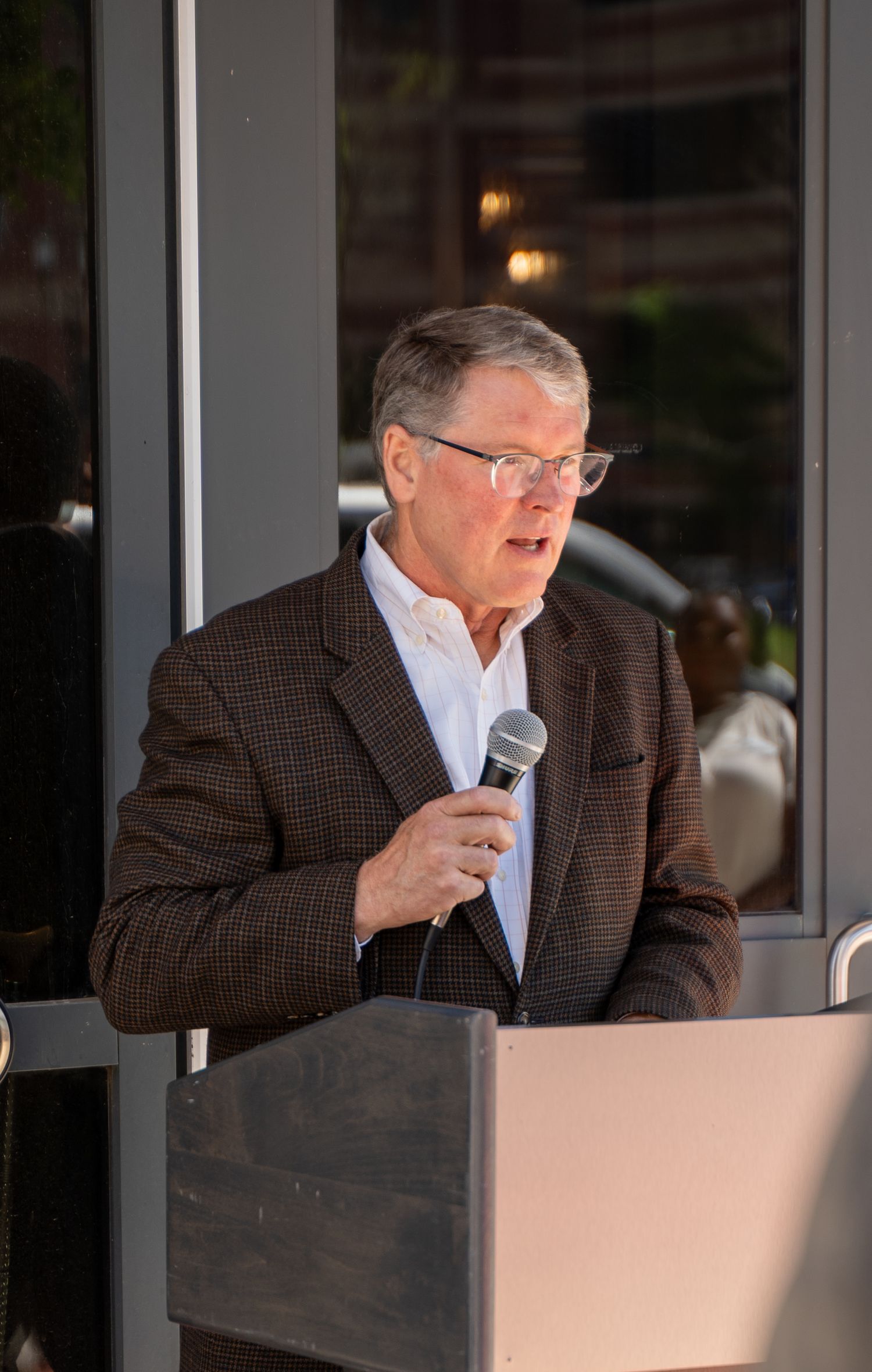 An older man in a brown blazer speaks at a podium while holding a microphone at a formal event.