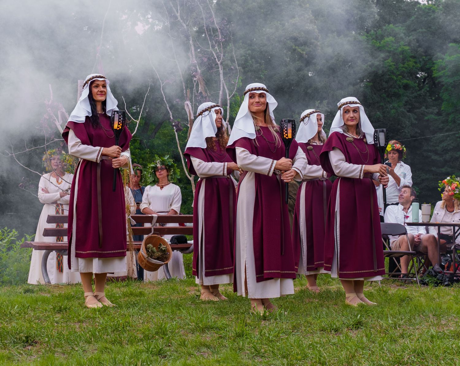 Women in traditional burgundy folk costumes perform a cultural dance outdoors amid misty green scenery.