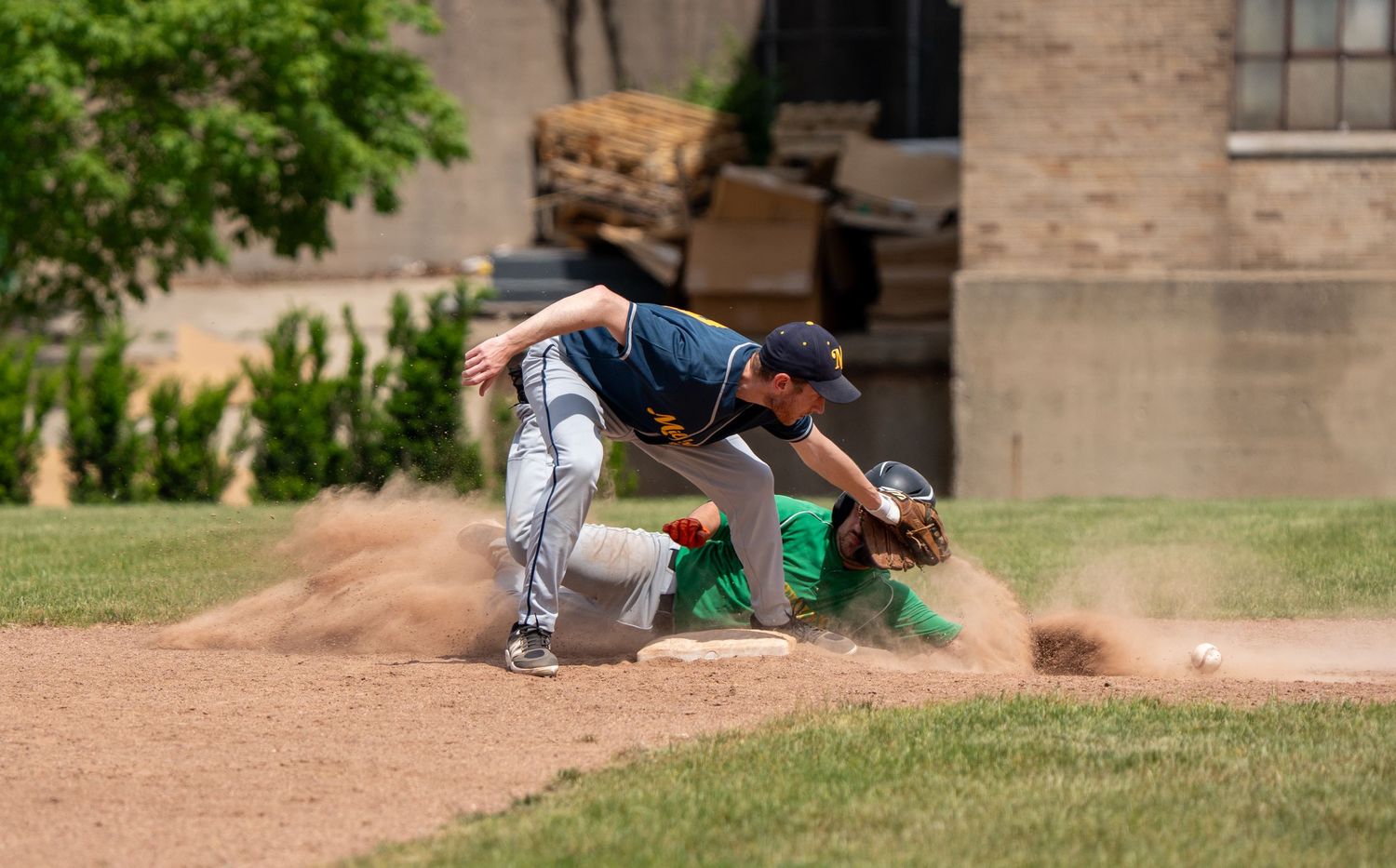 Baseball player slides into base in cloud of dust as fielder attempts tag during sunny outdoor game.