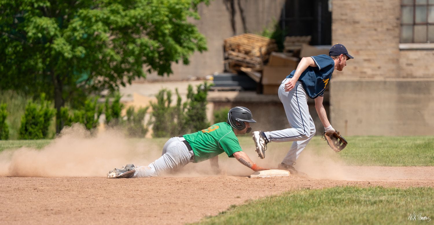 Baseball player in green slides into base as fielder in blue catches ball during outdoor game.