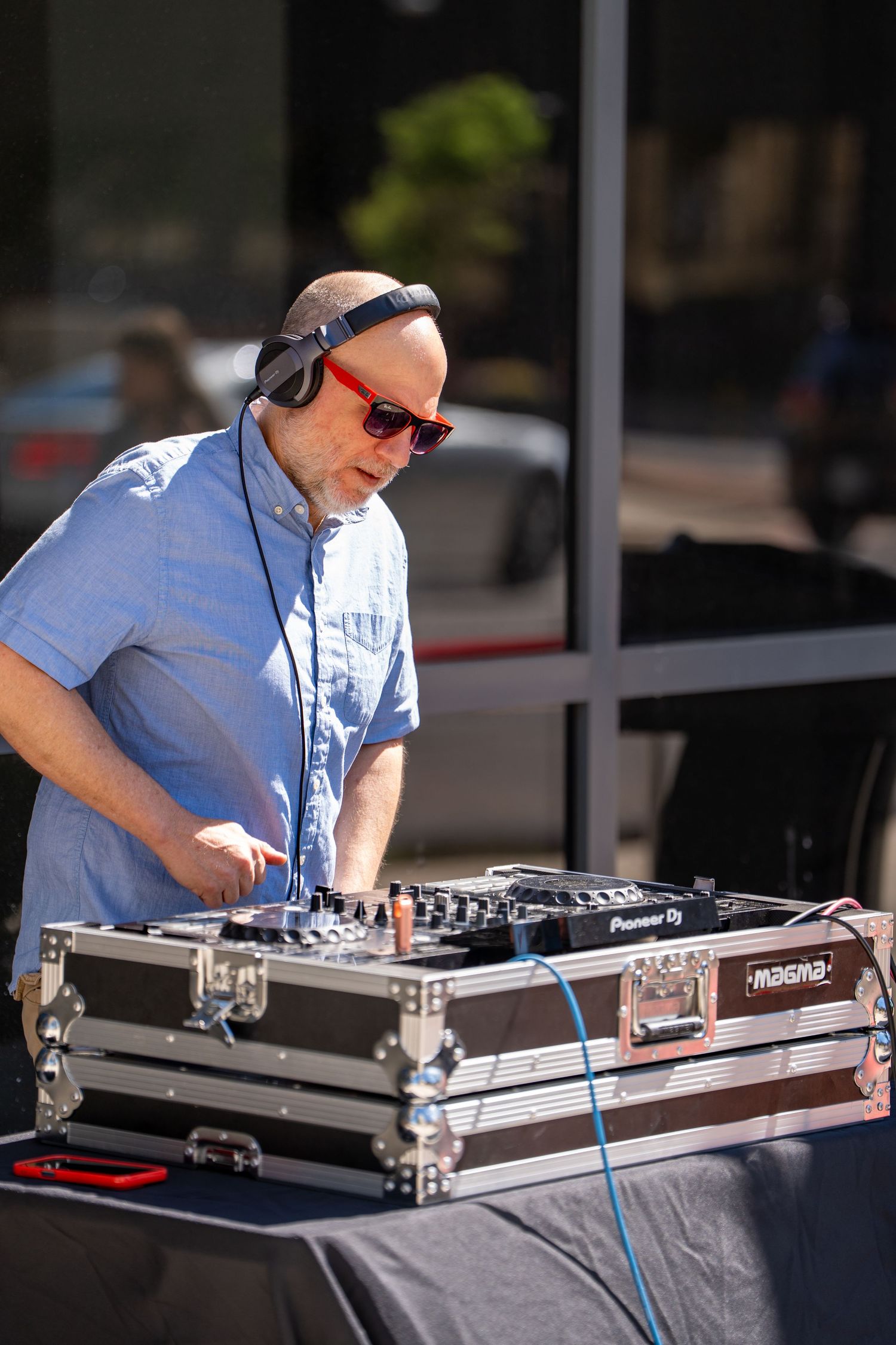Bald DJ wearing red sunglasses and headphones mixing music on a Pioneer DJ controller outdoors in sunlight.