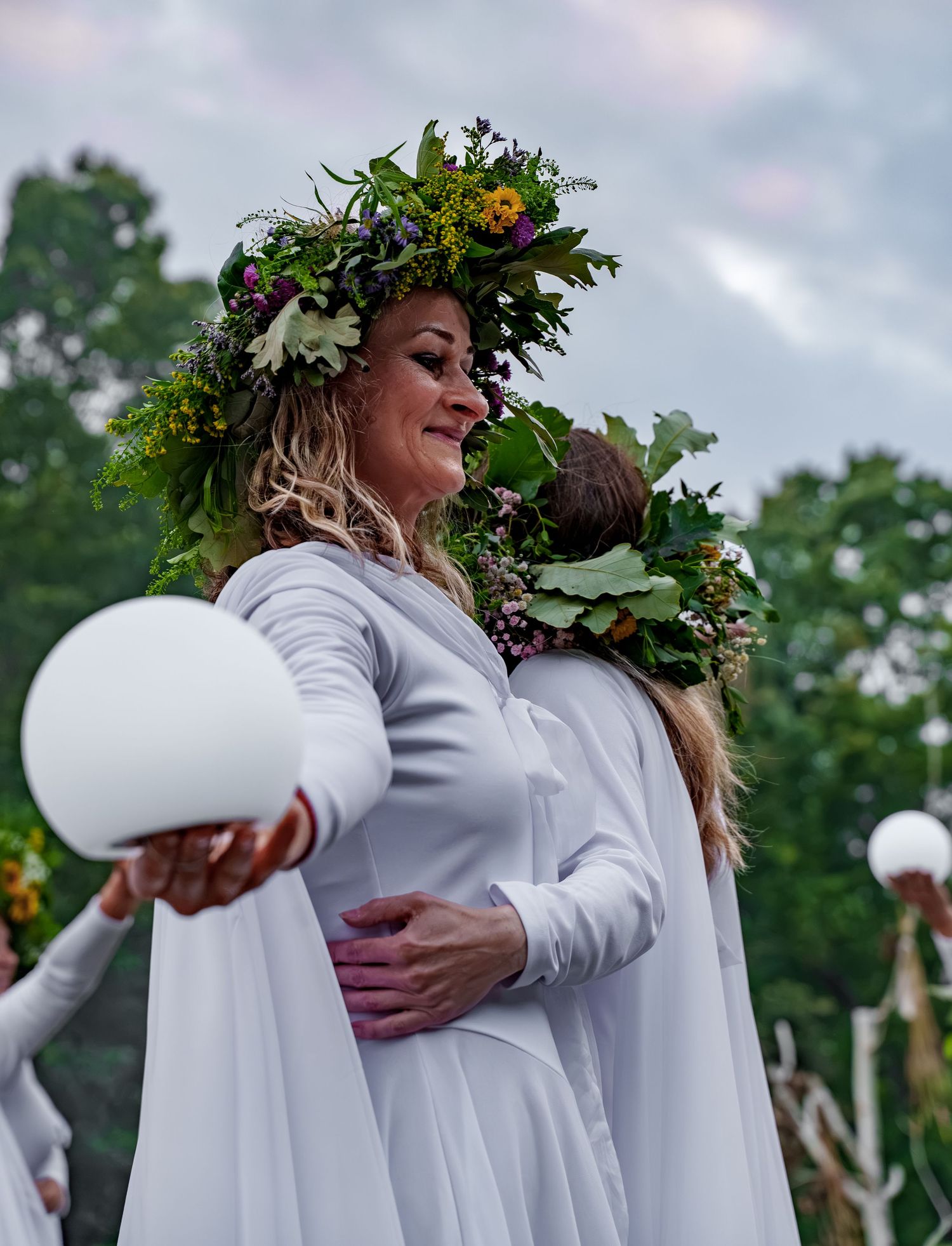 Woman in white dress wearing floral crown holds white orb at outdoor midsummer celebration.