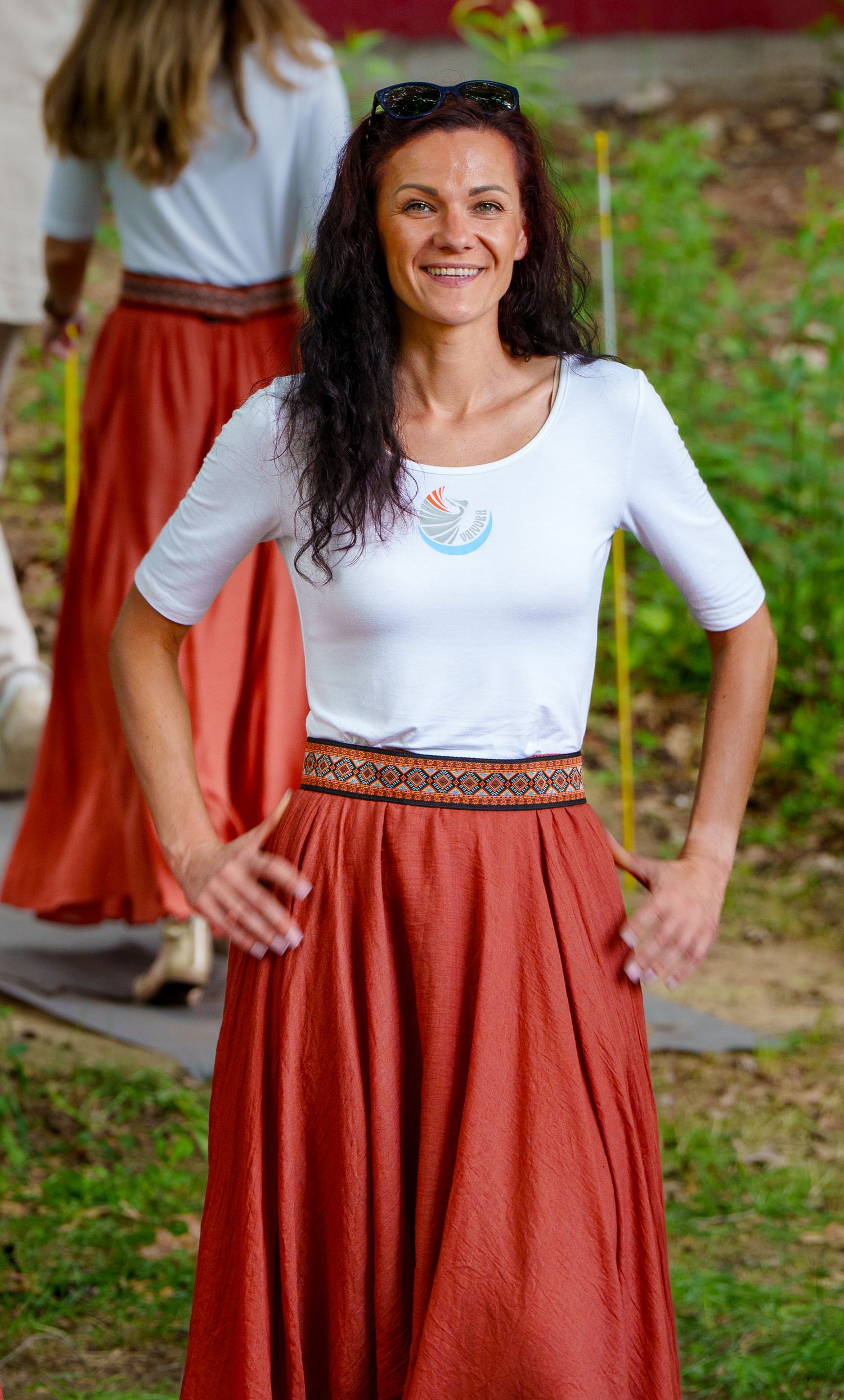 Smiling woman in white top and rust-colored maxi skirt with decorative belt poses outdoors.