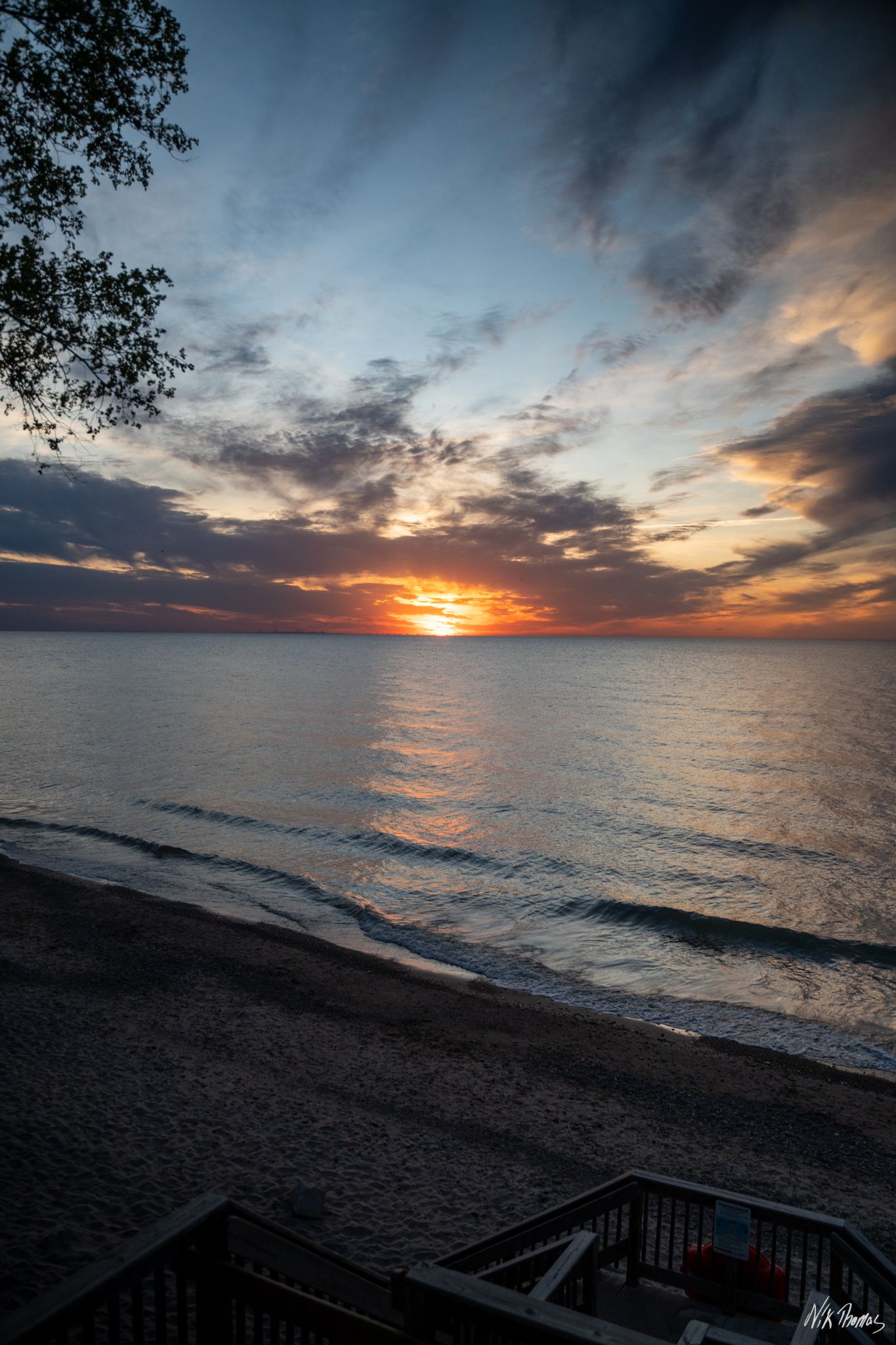 Stunning sunset over calm lake waters with dramatic clouds reflecting orange and blue hues on a sandy beach.
