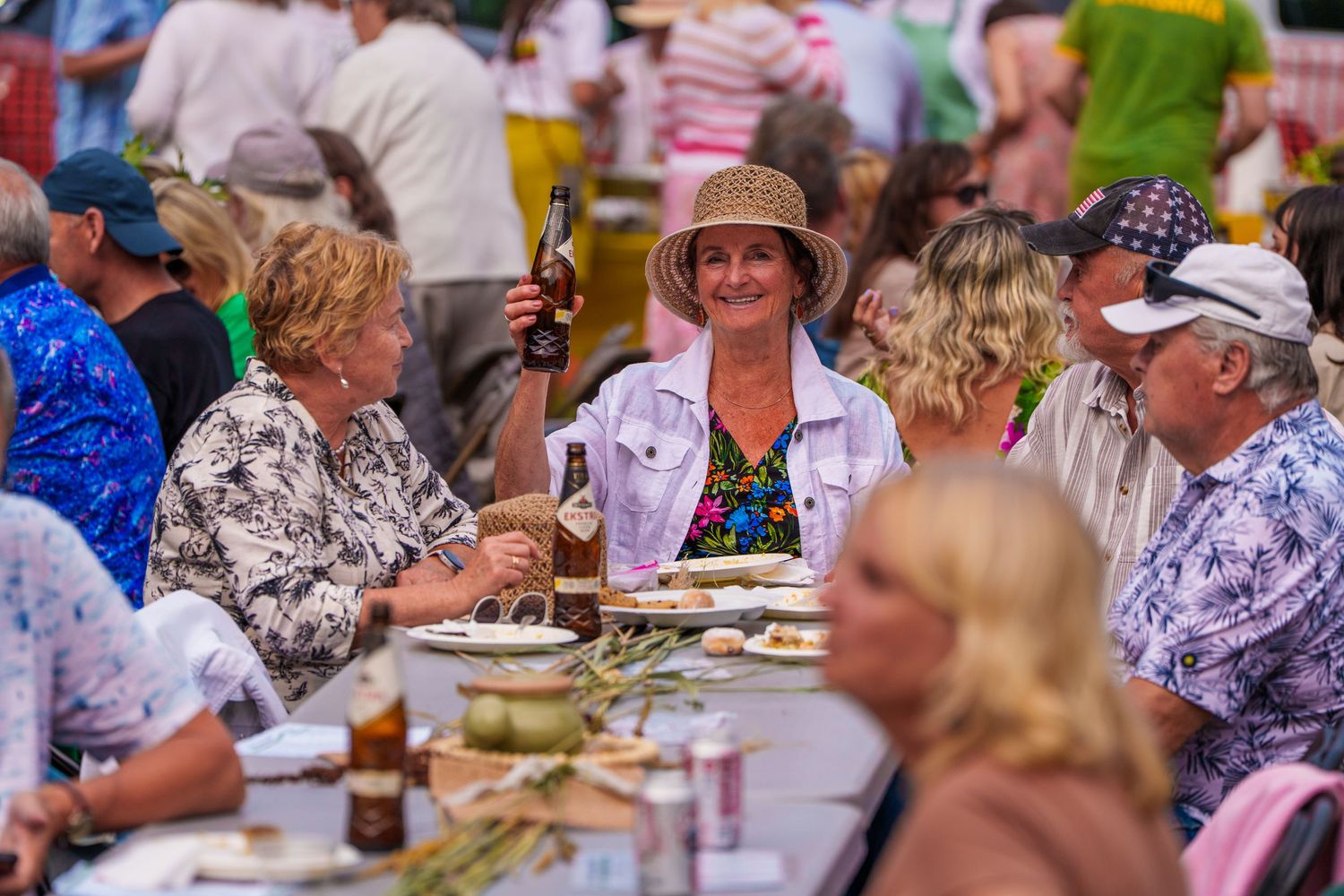 Woman holding bottle smiling at crowded outdoor festival dining table in summer.