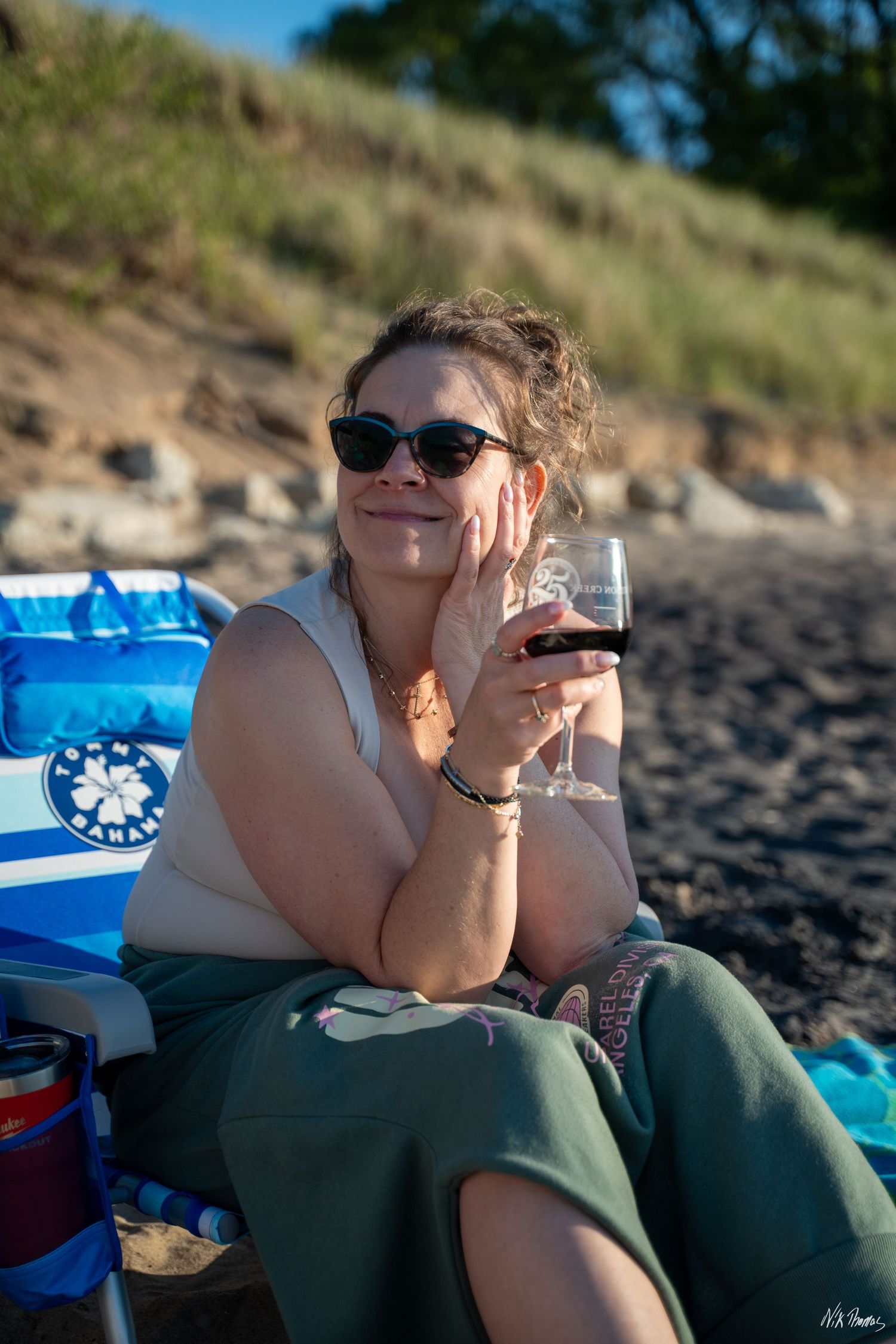 Woman relaxing on beach chair holding red wine glass, wearing sunglasses, smiling outdoors near sandy dunes.
