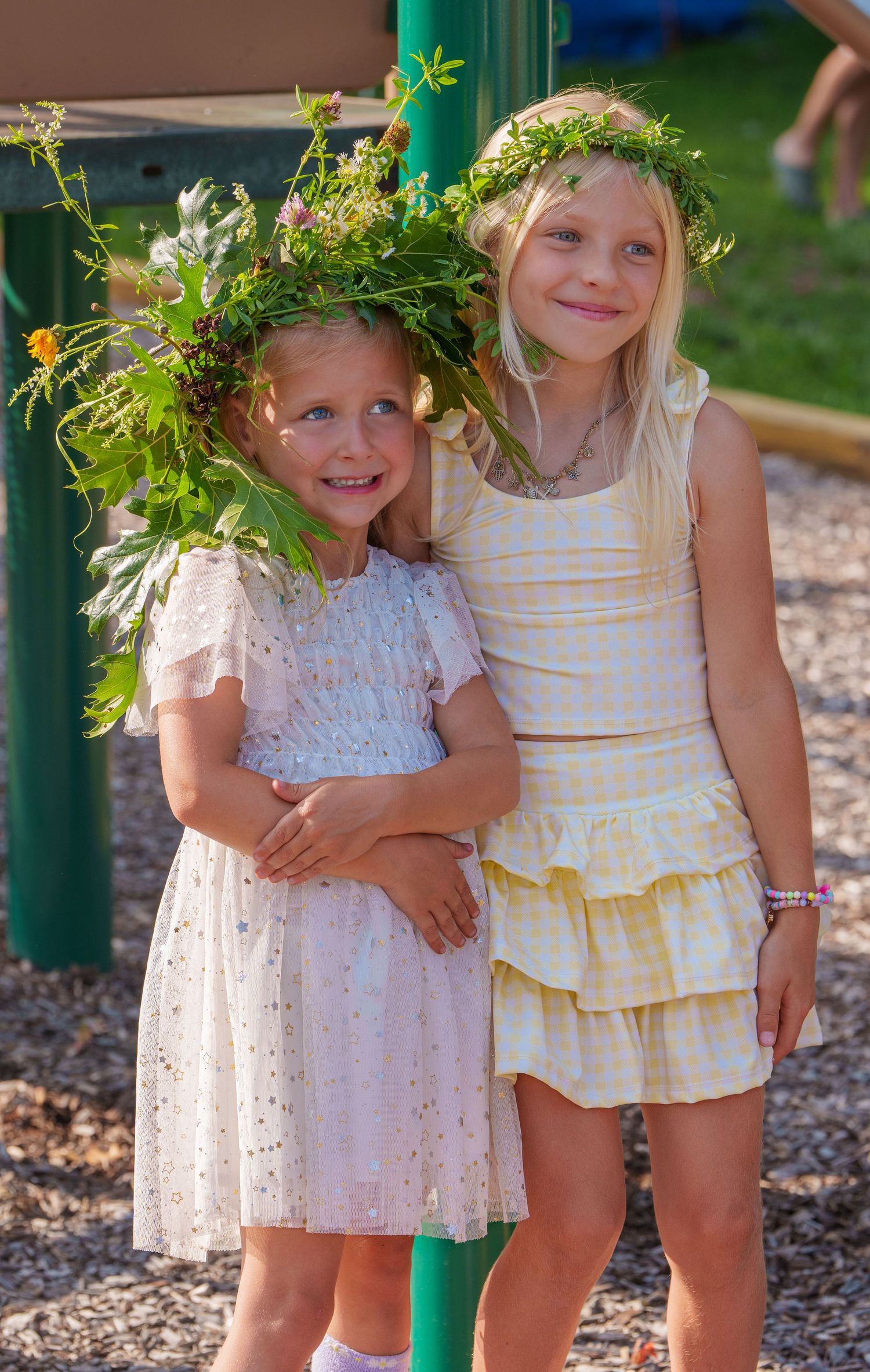 Two young girls wearing flower crowns in summer dresses smiling together at an outdoor playground.