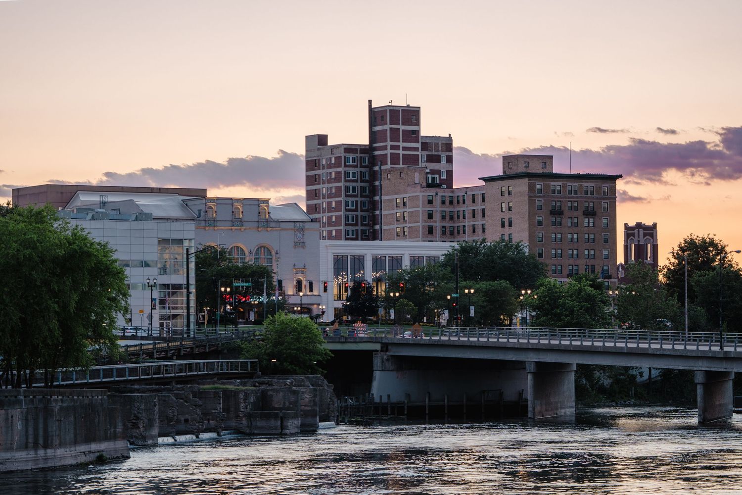Downtown cityscape at dusk with a river and bridge in the foreground, featuring historic brick buildings under a colorful sky.