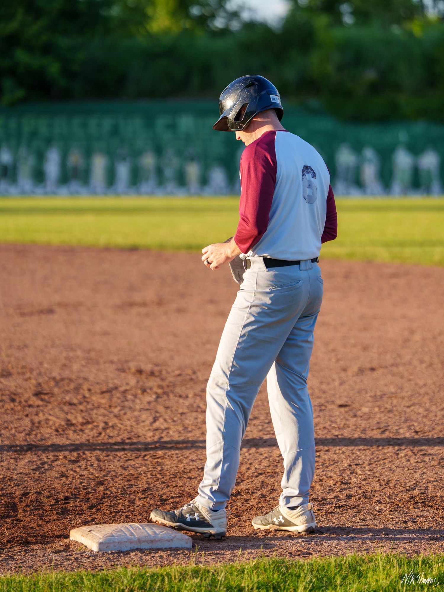 A baseball player in maroon and white uniform stands on base wearing a black batting helmet during a game.