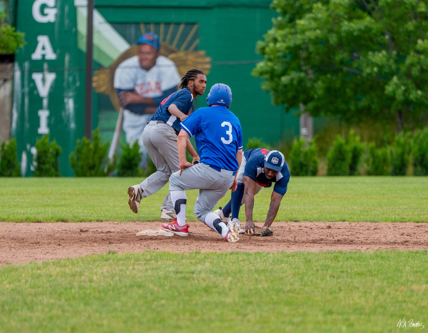 Baseball players in blue uniforms compete at second base during an outdoor game on a green grass field.