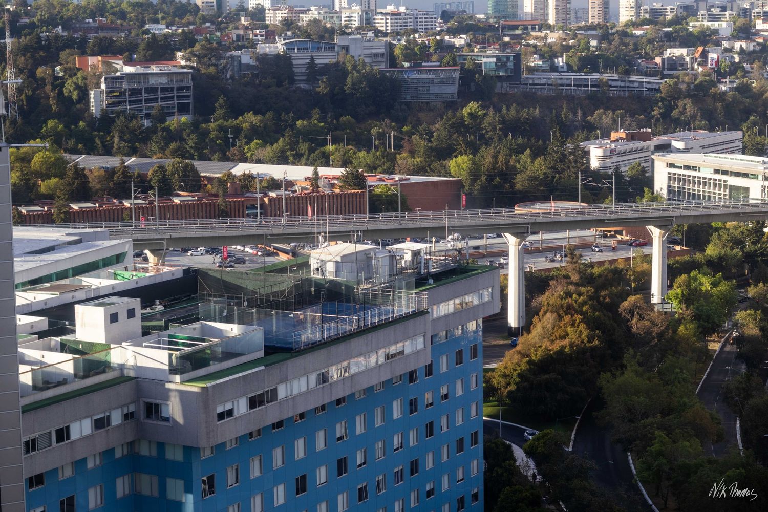 Aerial view of modern urban buildings with a rooftop terrace, elevated highway, and lush green trees in a city landscape.
