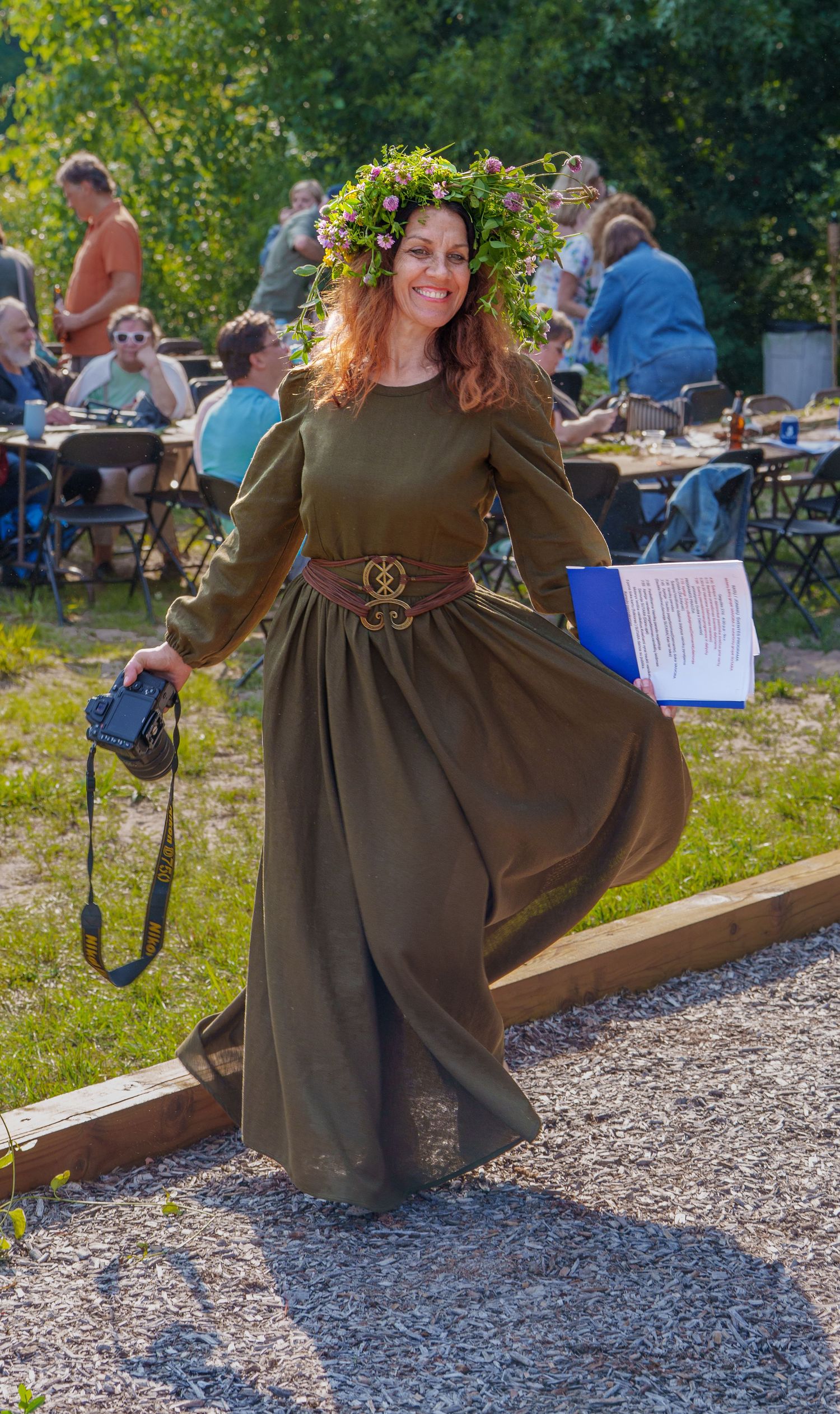 Woman in medieval green dress and floral crown smiles at outdoor Renaissance festival event.