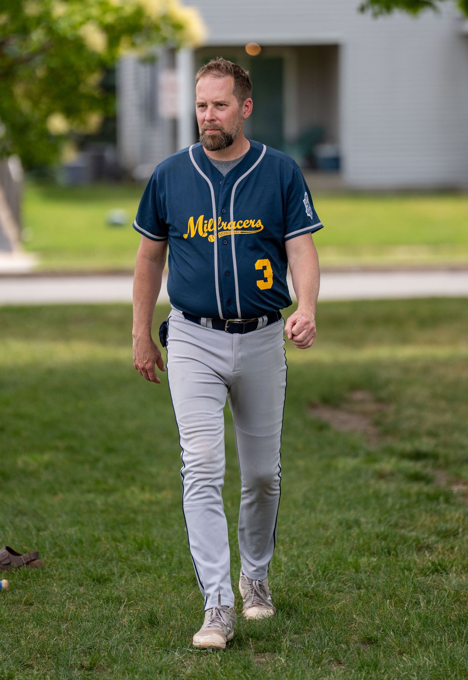 Man in navy Midtracers baseball jersey number 3 with gray pants walking on grass field.