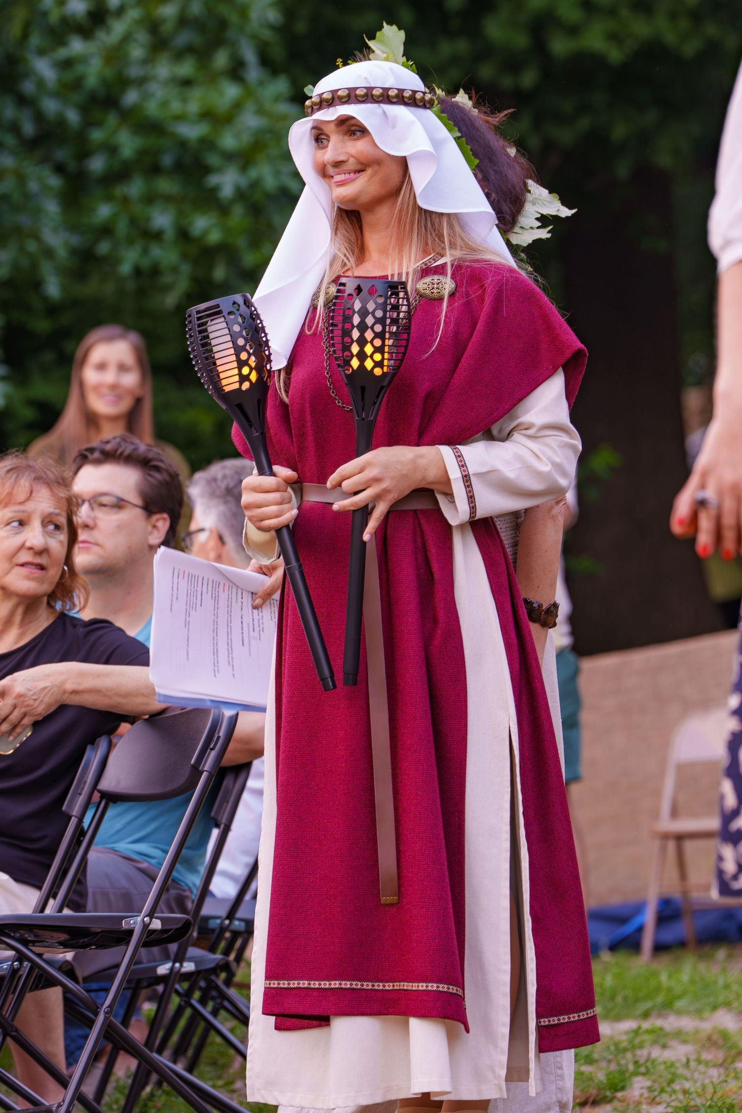 Woman in traditional Lithuanian costume holding torches at an outdoor cultural festival ceremony.