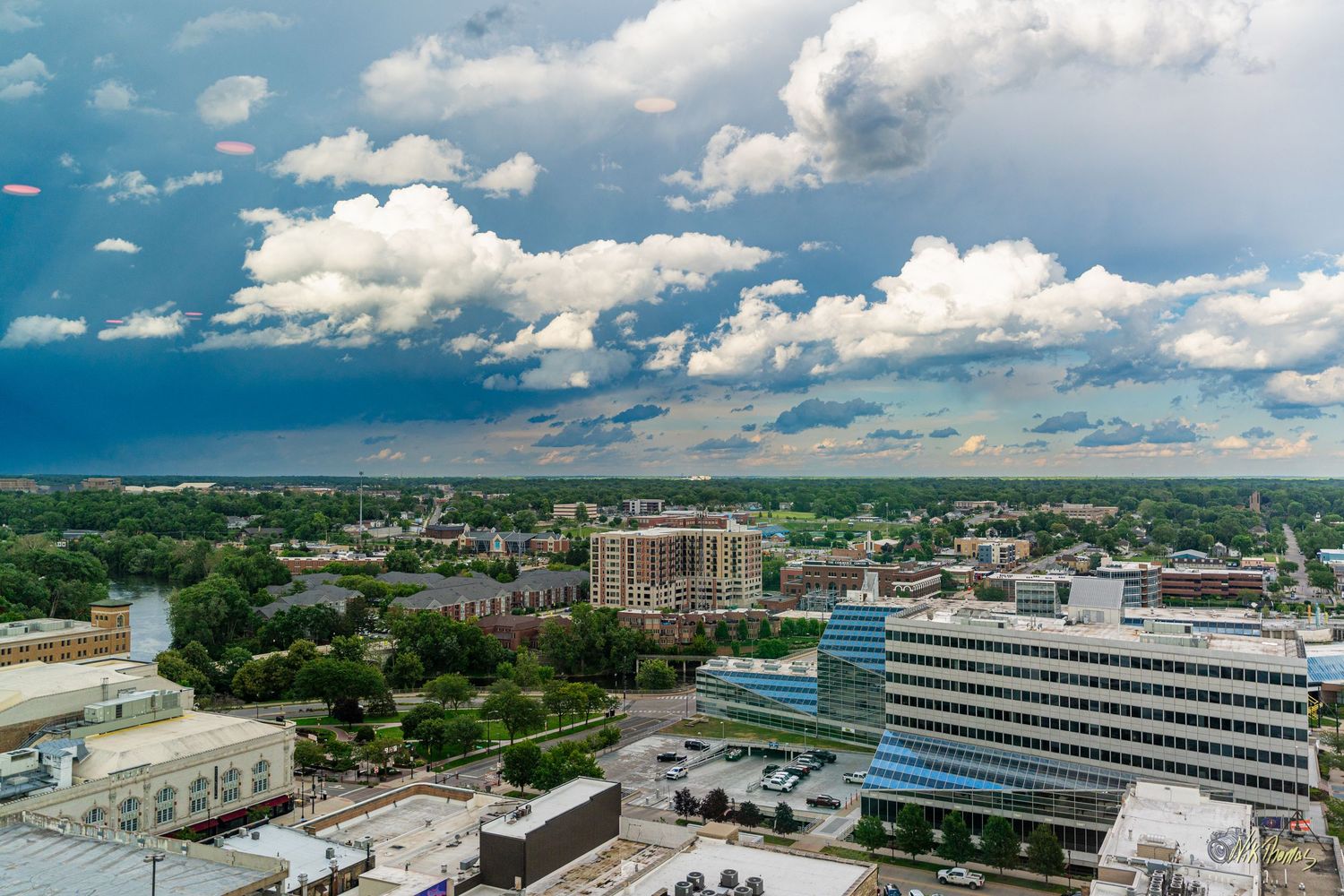 Aerial view of a mid-sized city skyline with lush green trees, buildings, and dramatic blue sky with white clouds.