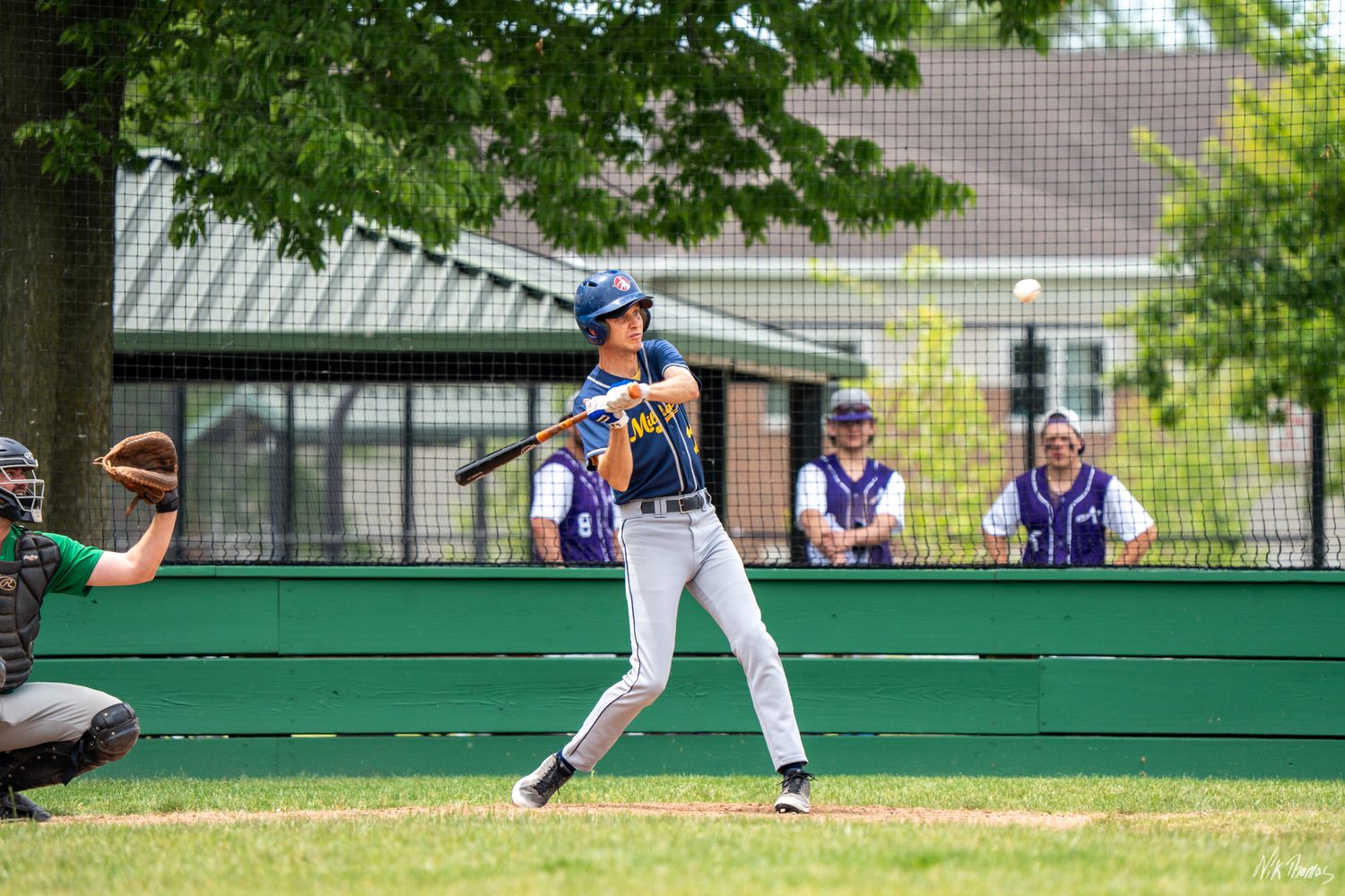Baseball batter in purple uniform swings at pitch during sunny outdoor game, with catcher and dugout players visible.