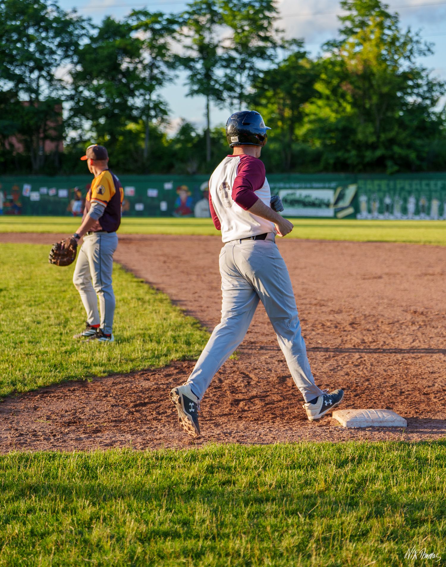 Baseball baserunner rounds first base while fielder stands nearby during an evening little league game.