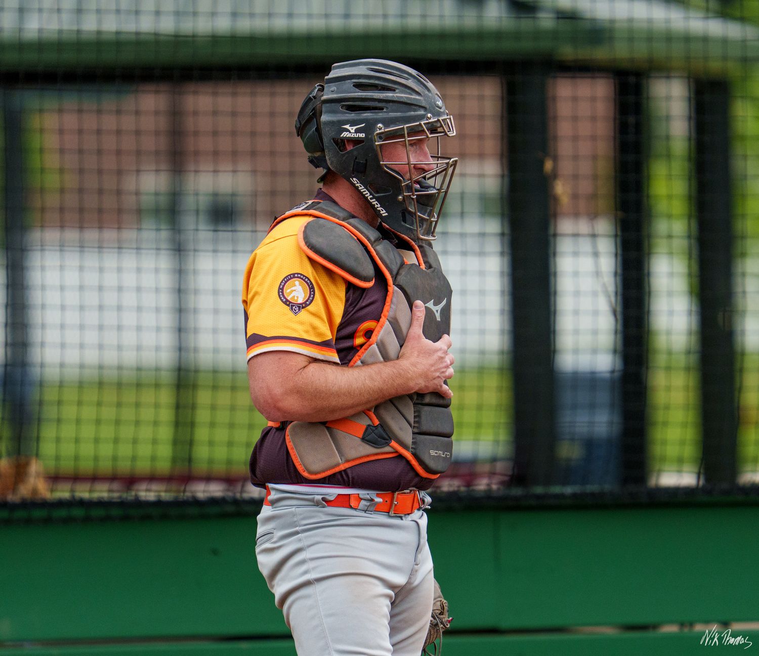 Baseball catcher in gold and red uniform wearing helmet and chest protector during outdoor game.
