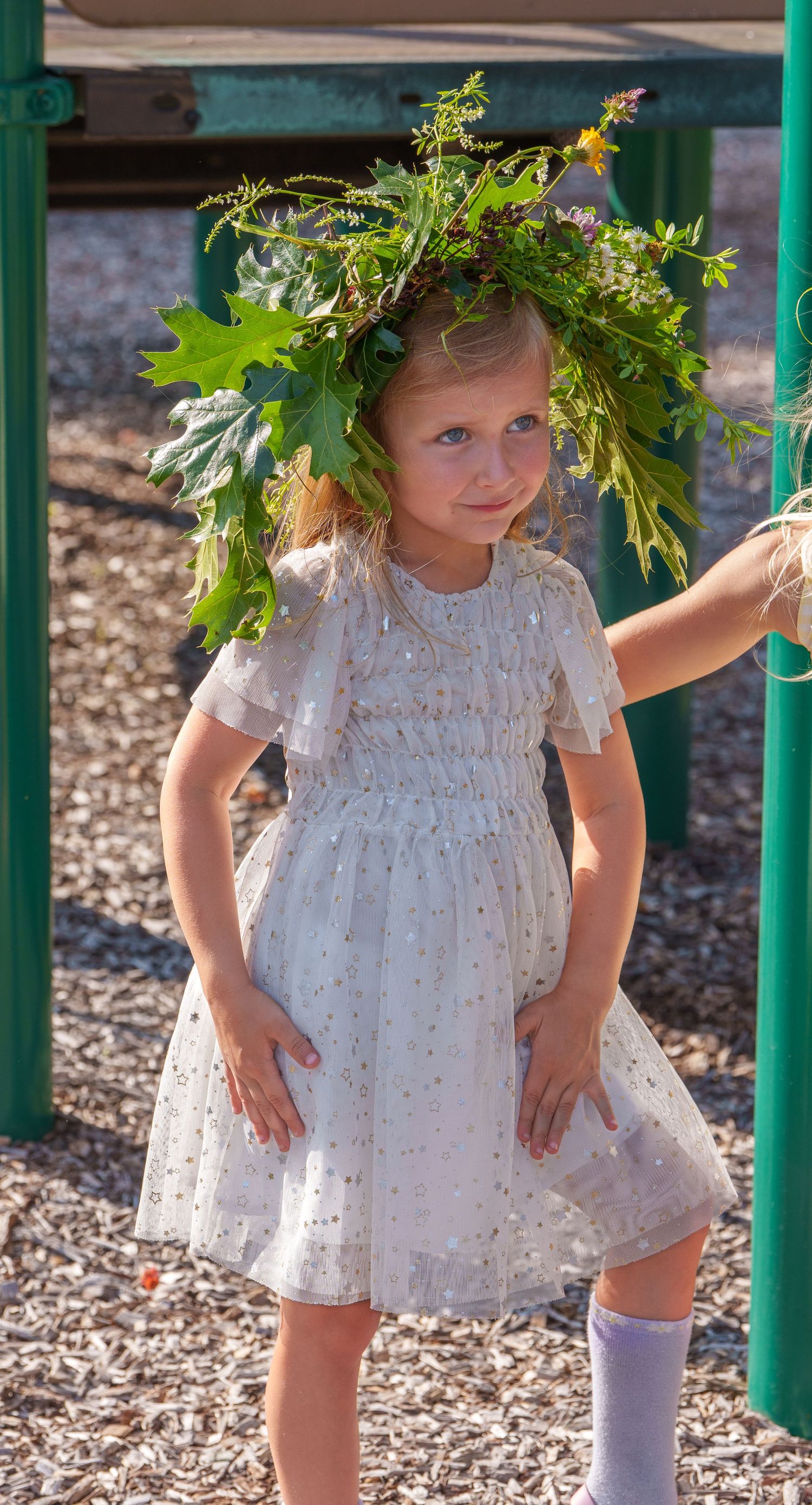 Young girl in a white dress wearing a large leafy green wreath crown at an outdoor playground.