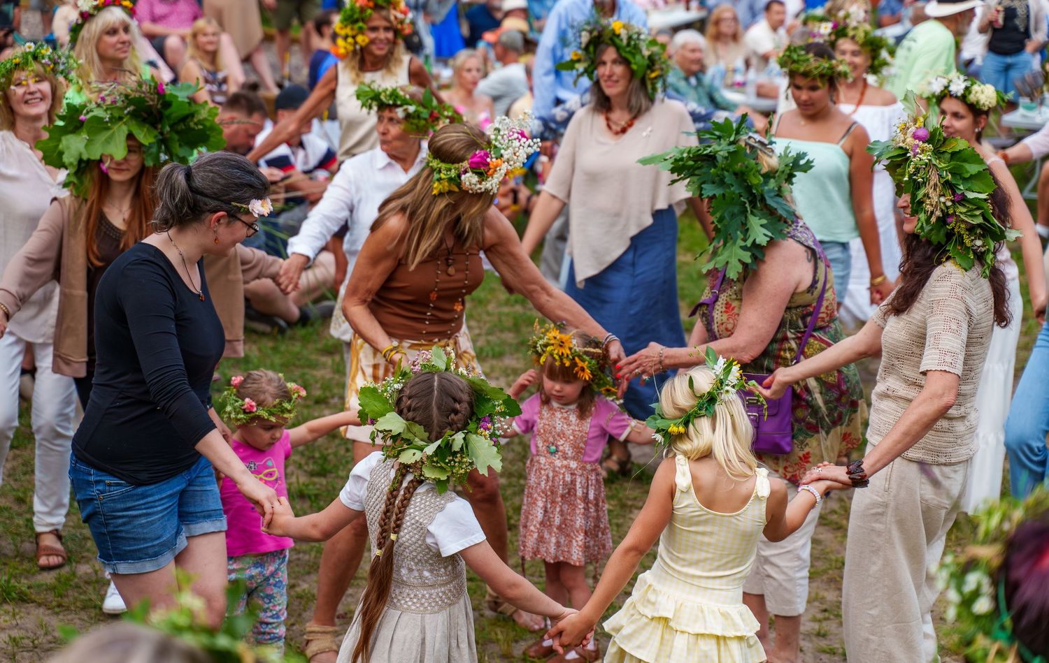 Women and children wearing flower crowns dance in a circle outdoors at a midsummer celebration.