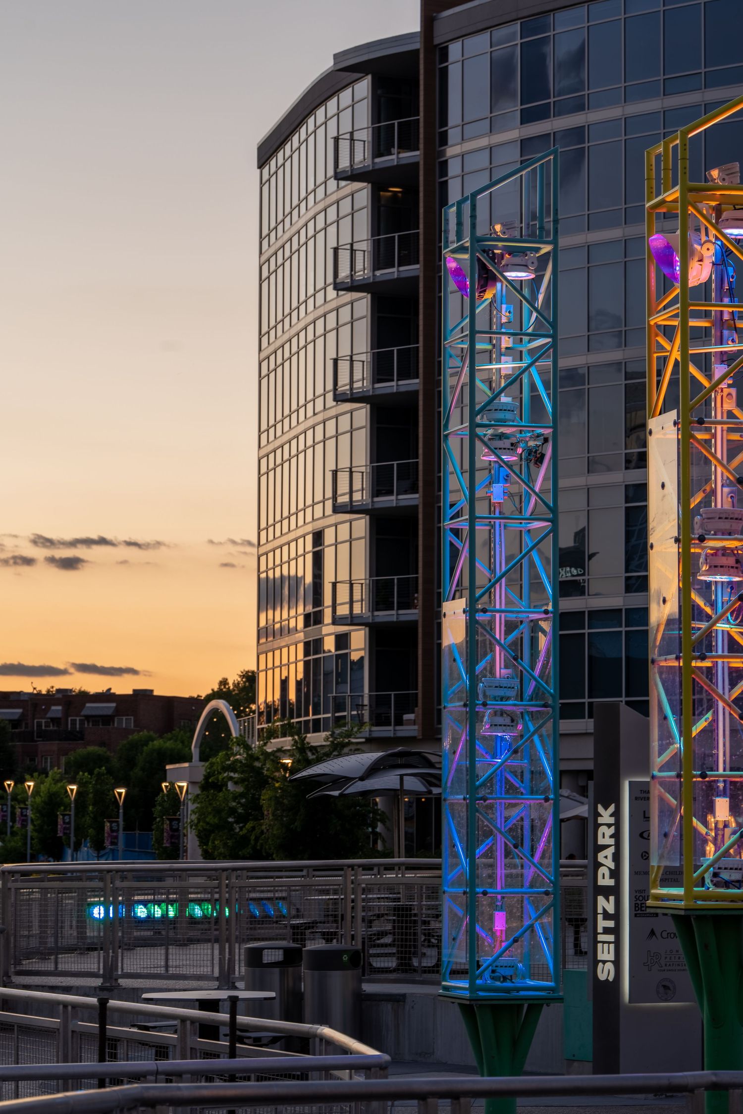 Colorful illuminated steel structures at Seitz Park glow at sunset beside a modern glass skyscraper in an urban cityscape.