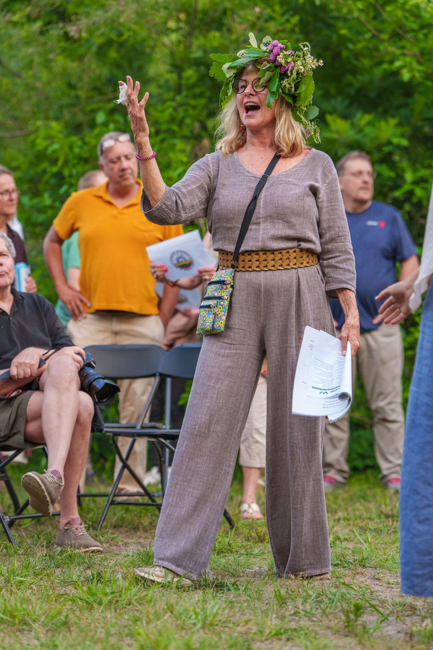 Woman in floral crown and gray jumpsuit gestures while speaking outdoors at a garden gathering.