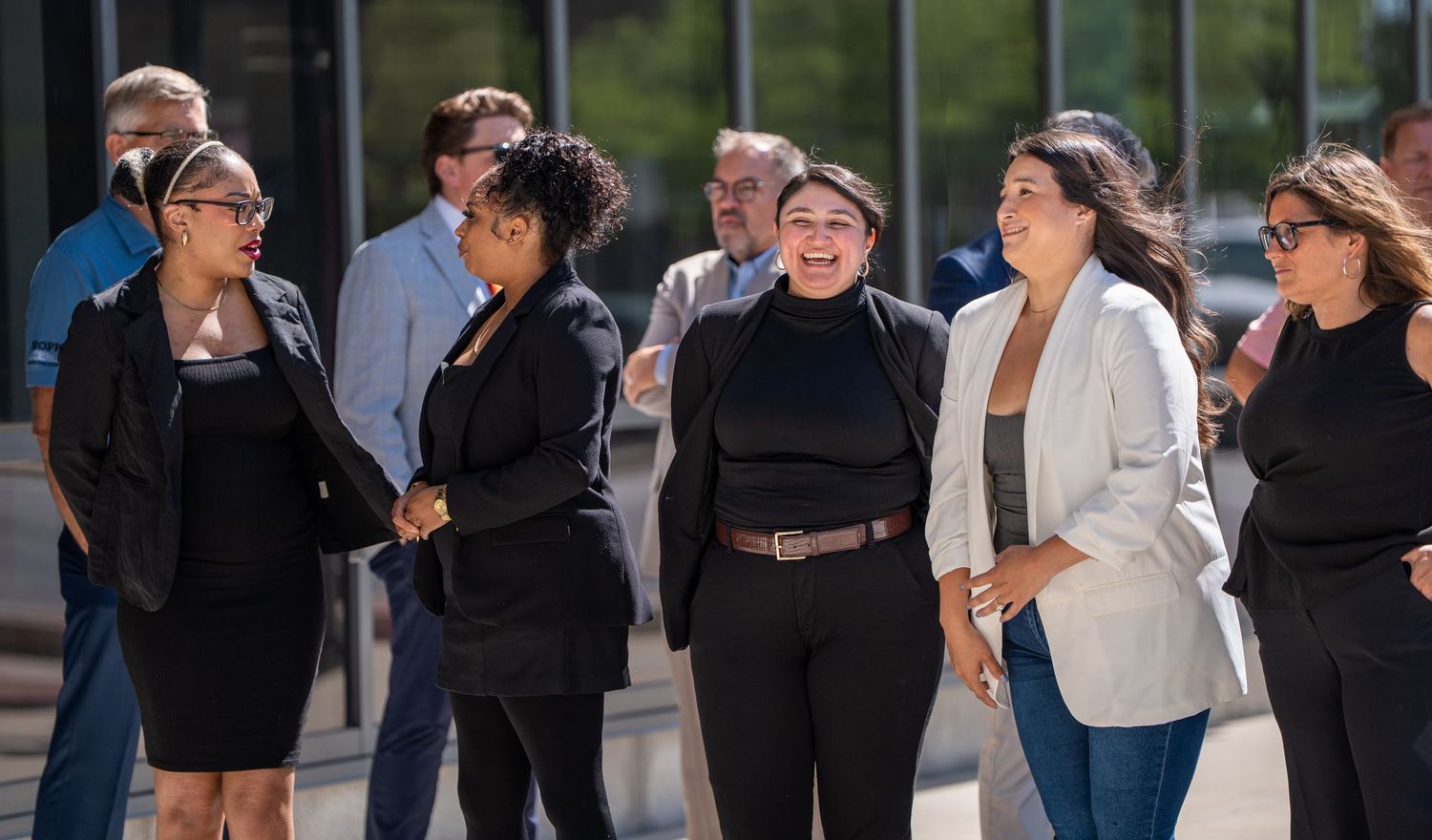 Diverse group of professionals in business attire networking outdoors near a modern building on a sunny day.