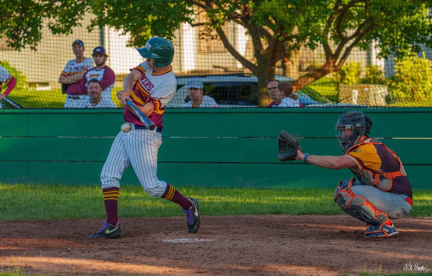 Baseball batter swings at pitch as catcher crouches during an outdoor evening game.