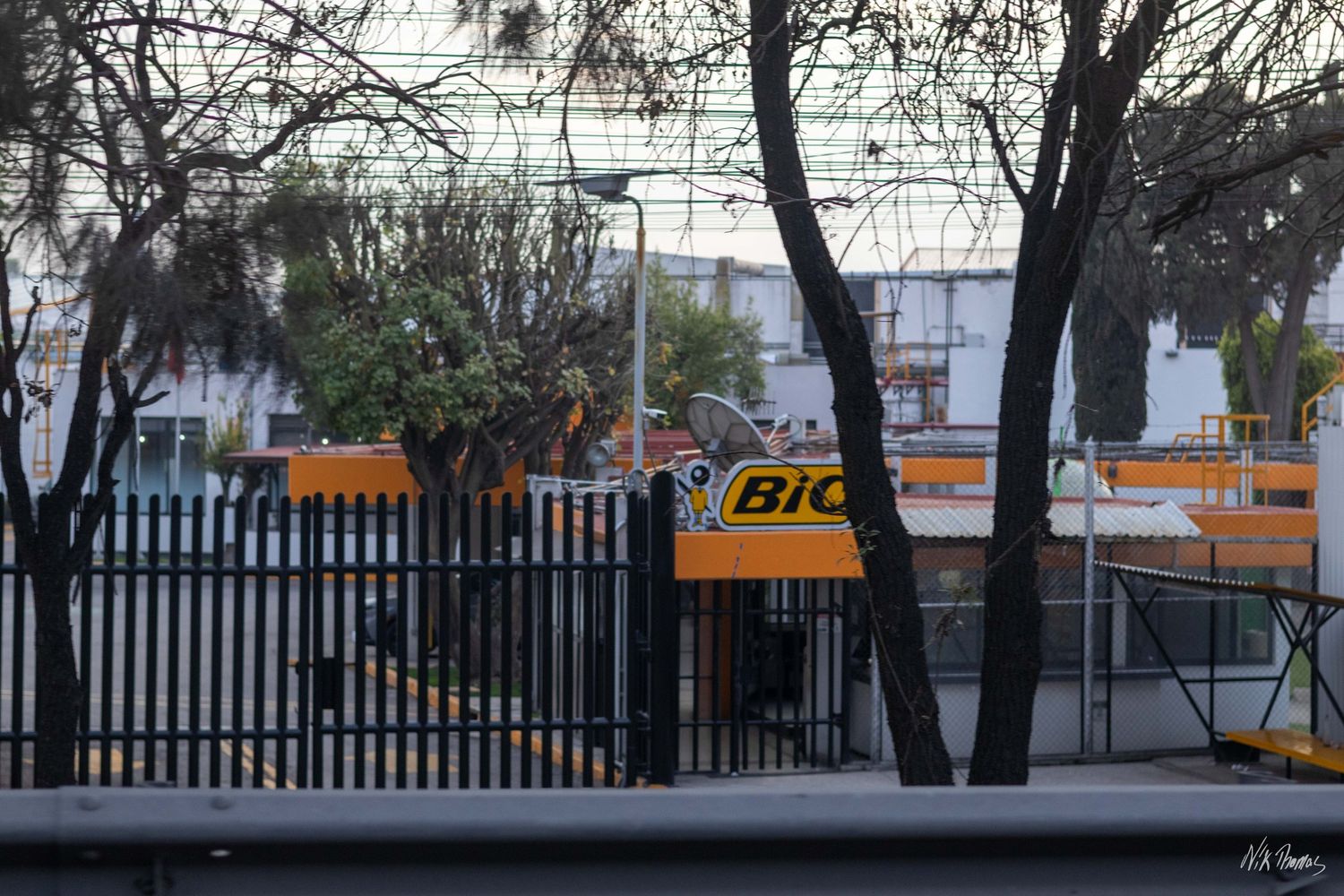 BIC branded yellow signage visible through black iron fence with bare trees in urban setting.