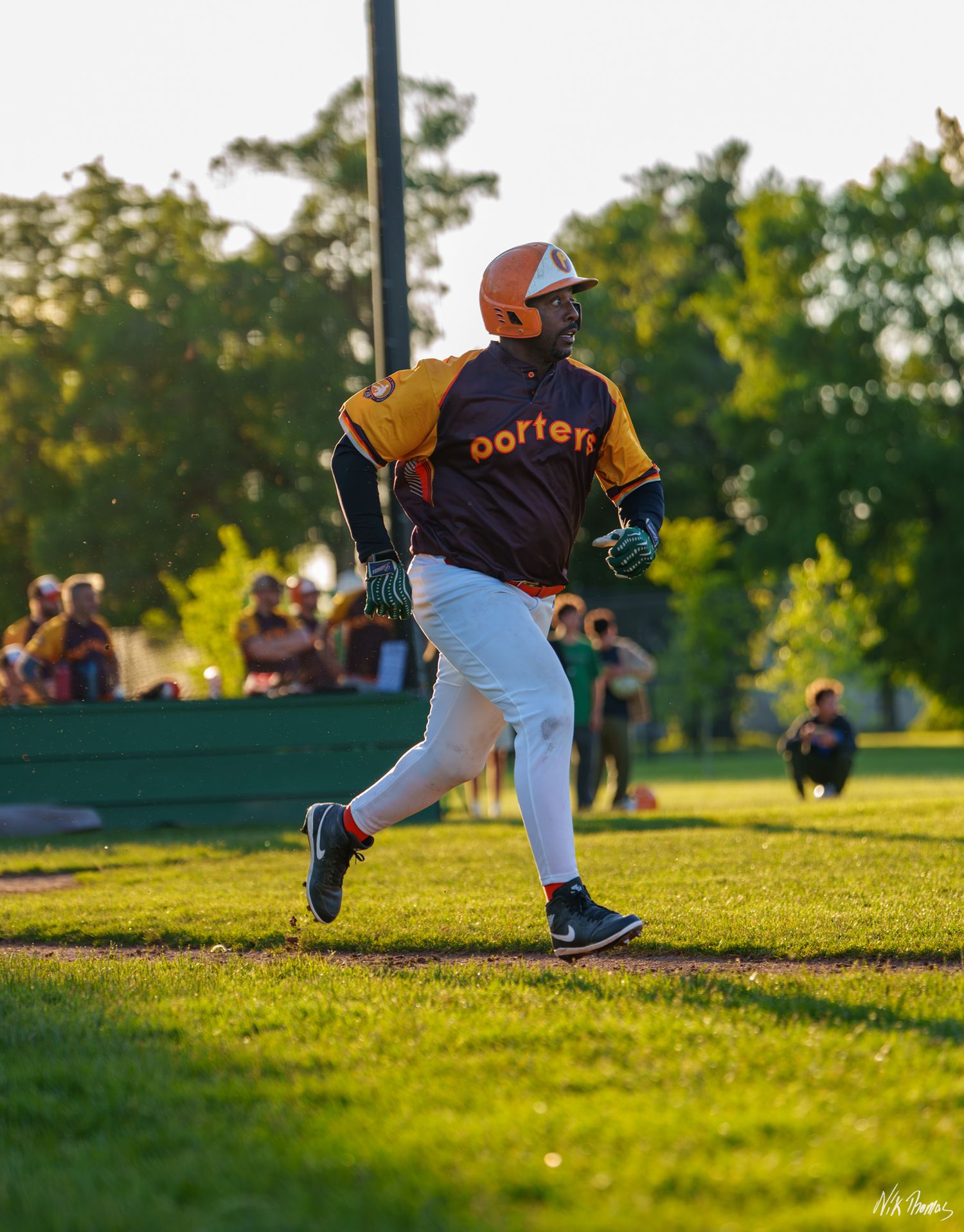 Baseball player in orange helmet and Oorten jersey runs bases during a sunny outdoor game with spectators in background.