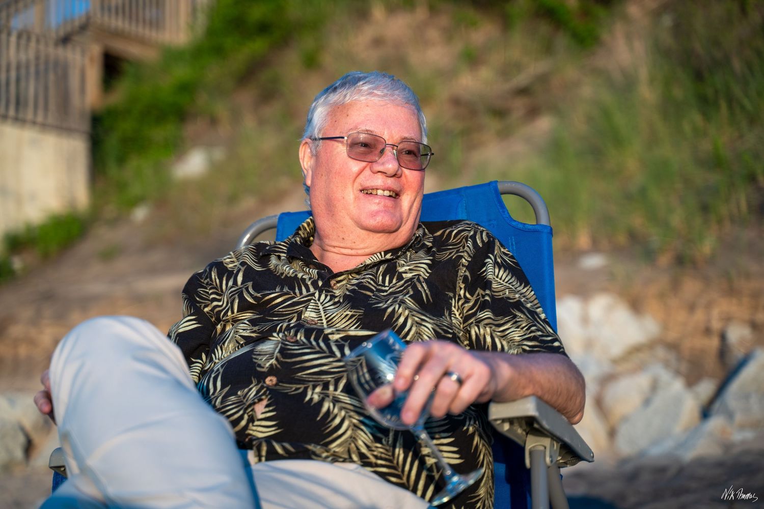 Smiling older man relaxing in blue beach chair wearing floral shirt and sunglasses holding a drink.