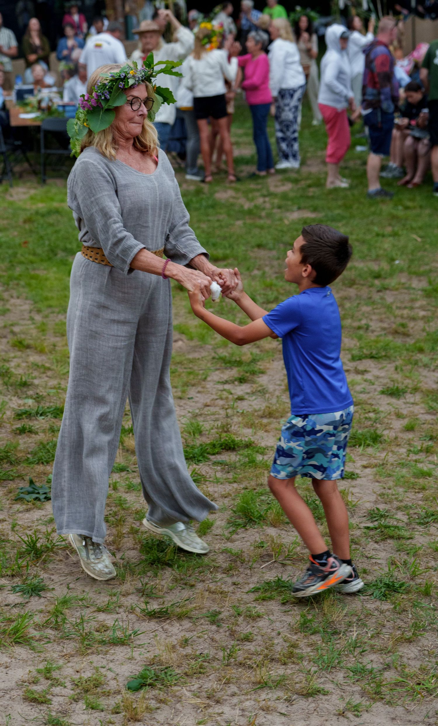 Woman wearing floral crown in gray outfit dances with young boy on grass at festival.