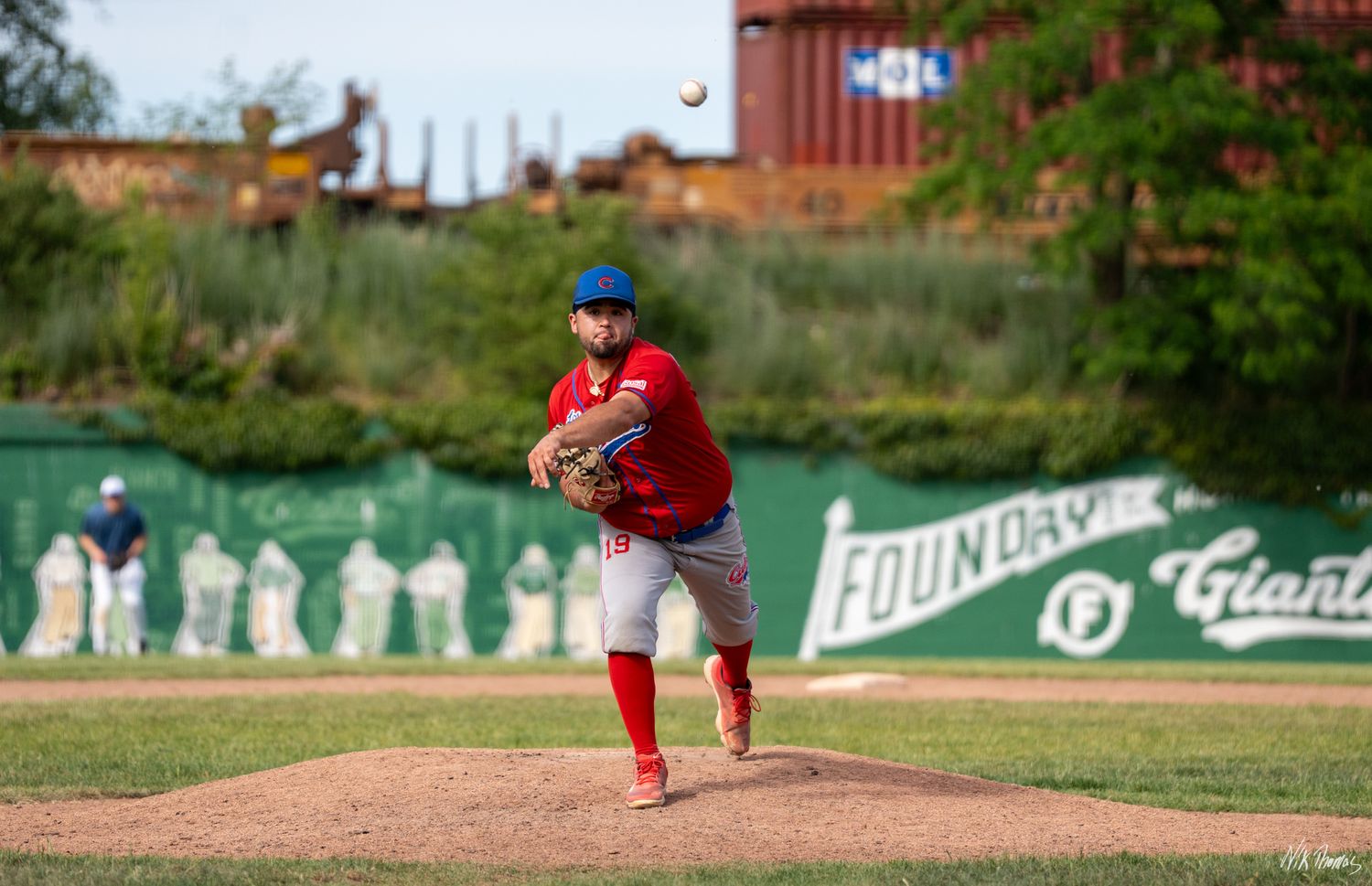 Baseball pitcher in red uniform mid-throw on a green field during a sunny evening game.